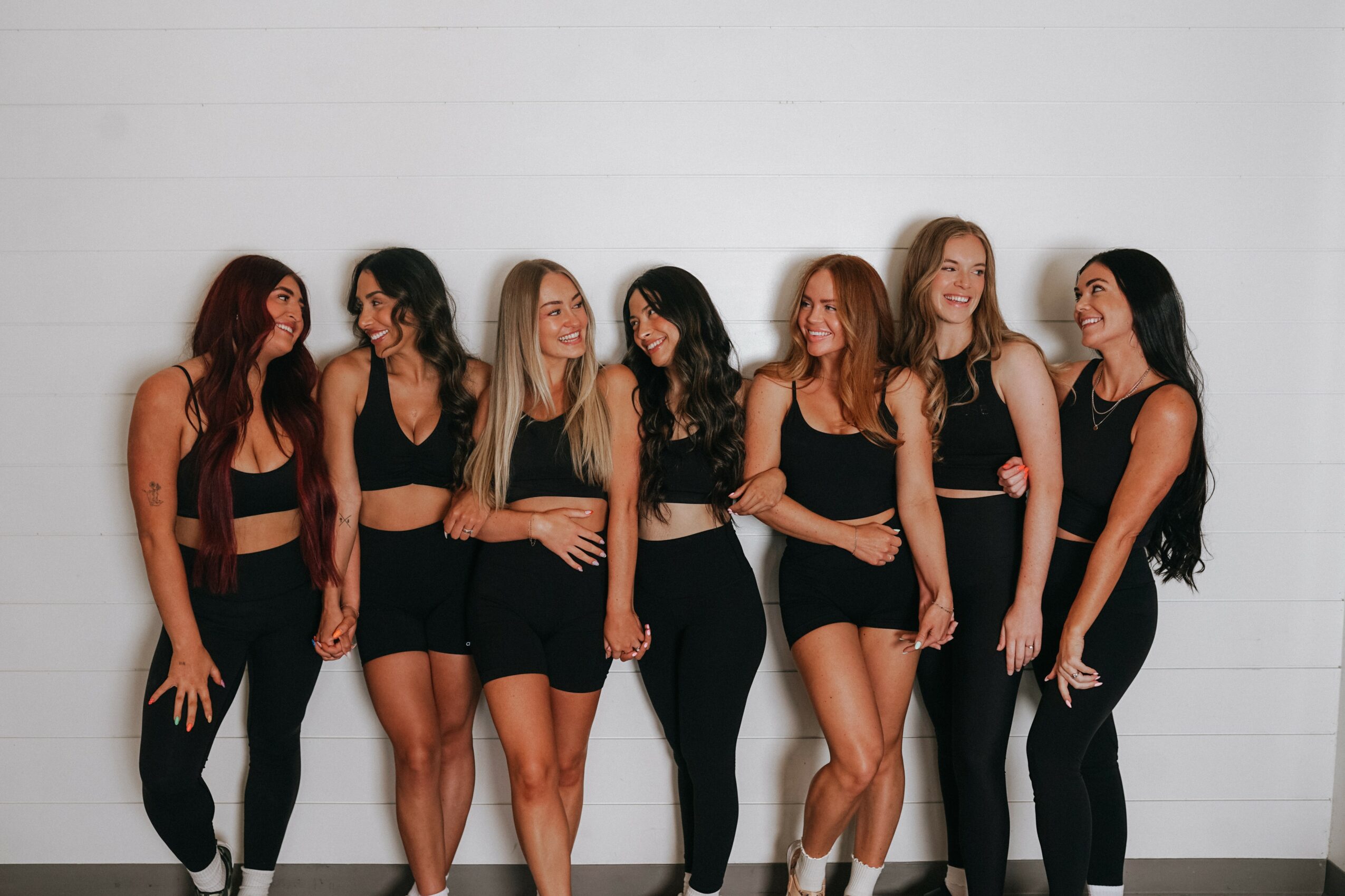 Seven women standing against a white wall, smiling and talking, wearing black outfits, in a casual group pose.