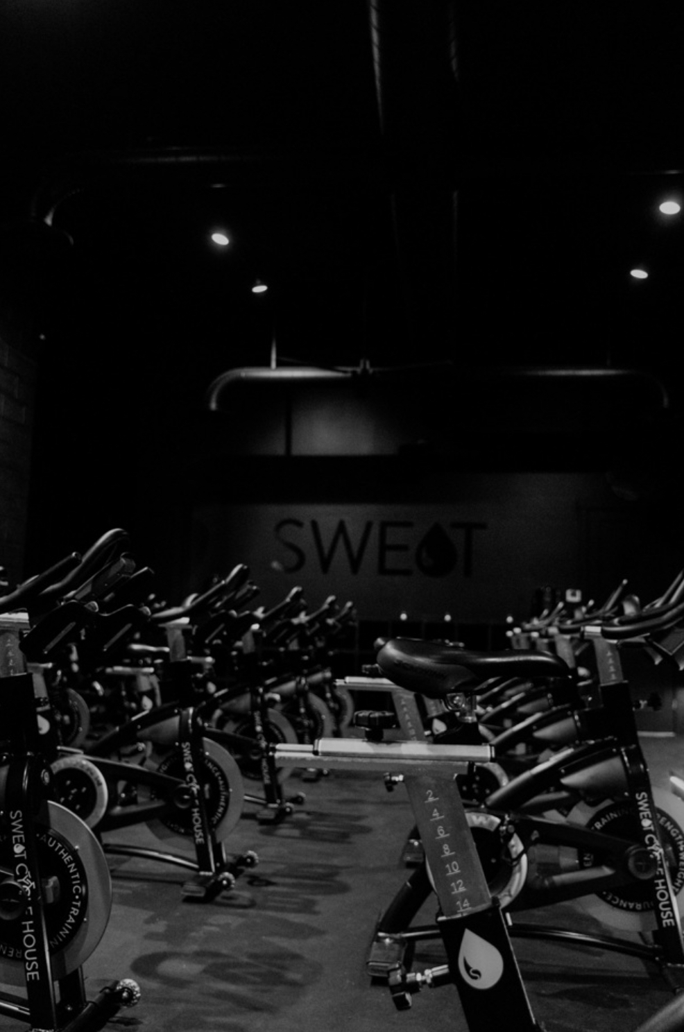 Indoor gym with exercise bikes and a sign reading 'SWEAT' in the background, dim lighting, and ceiling lights.