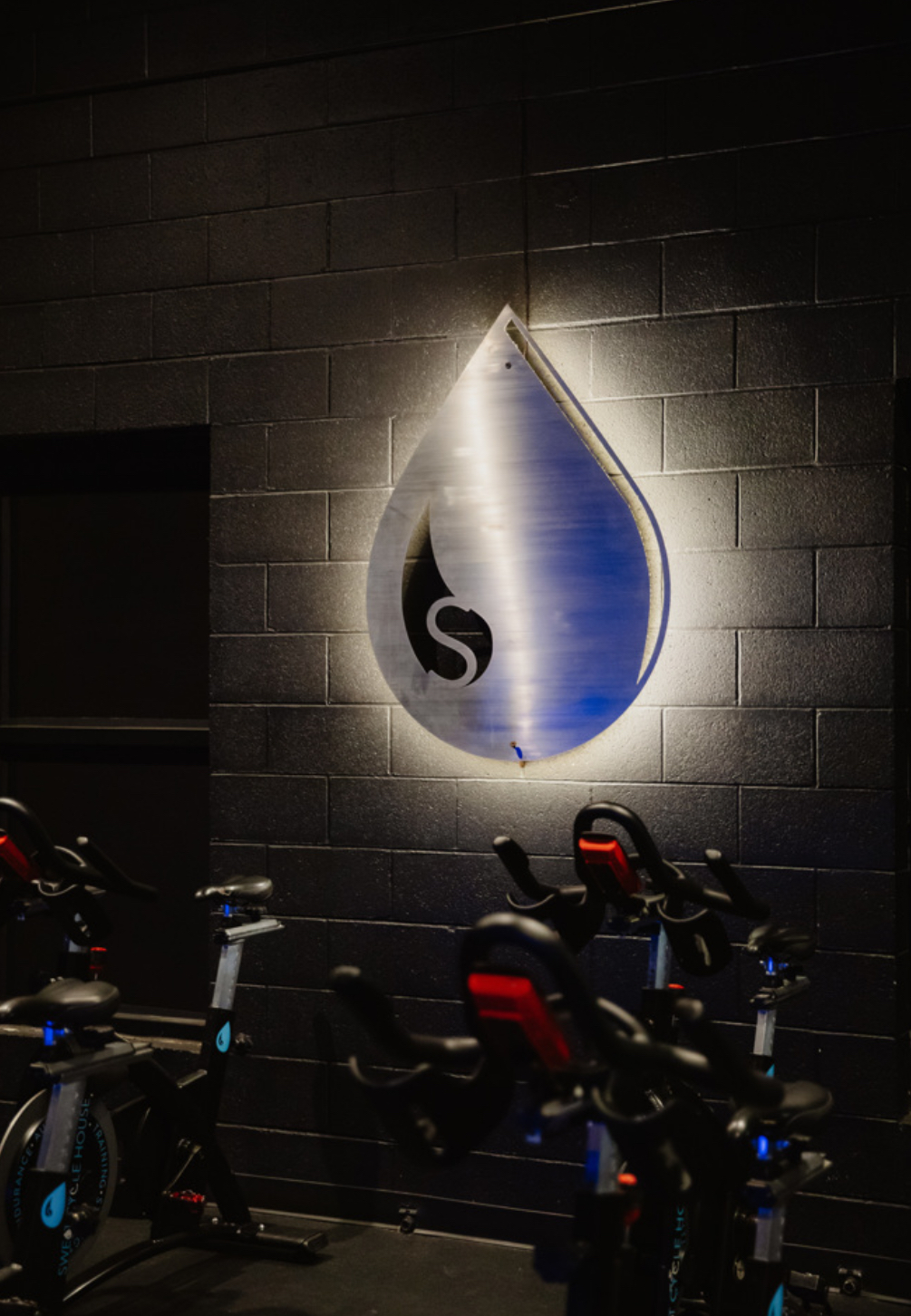 Backlit sign of a water droplet with a logo inside, mounted on a dark brick wall, bicycles in foreground.