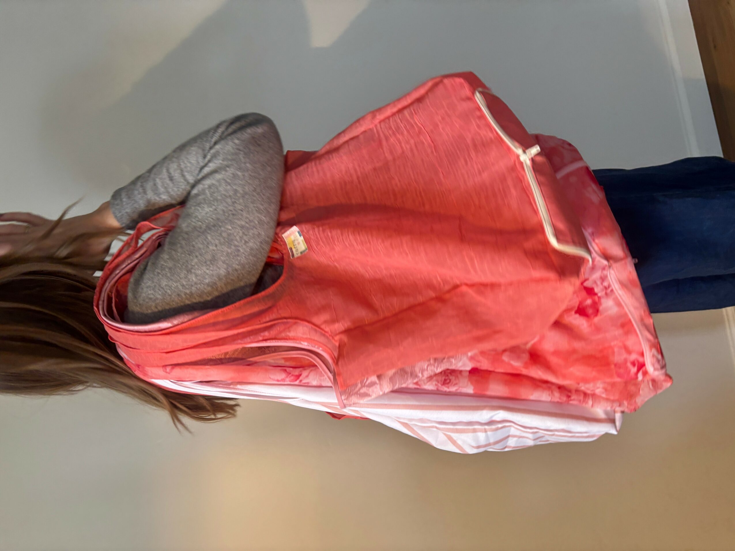 Child with long hair wearing a pink dress and gray long-sleeve shirt, standing sideways, arms crossed, indoors.