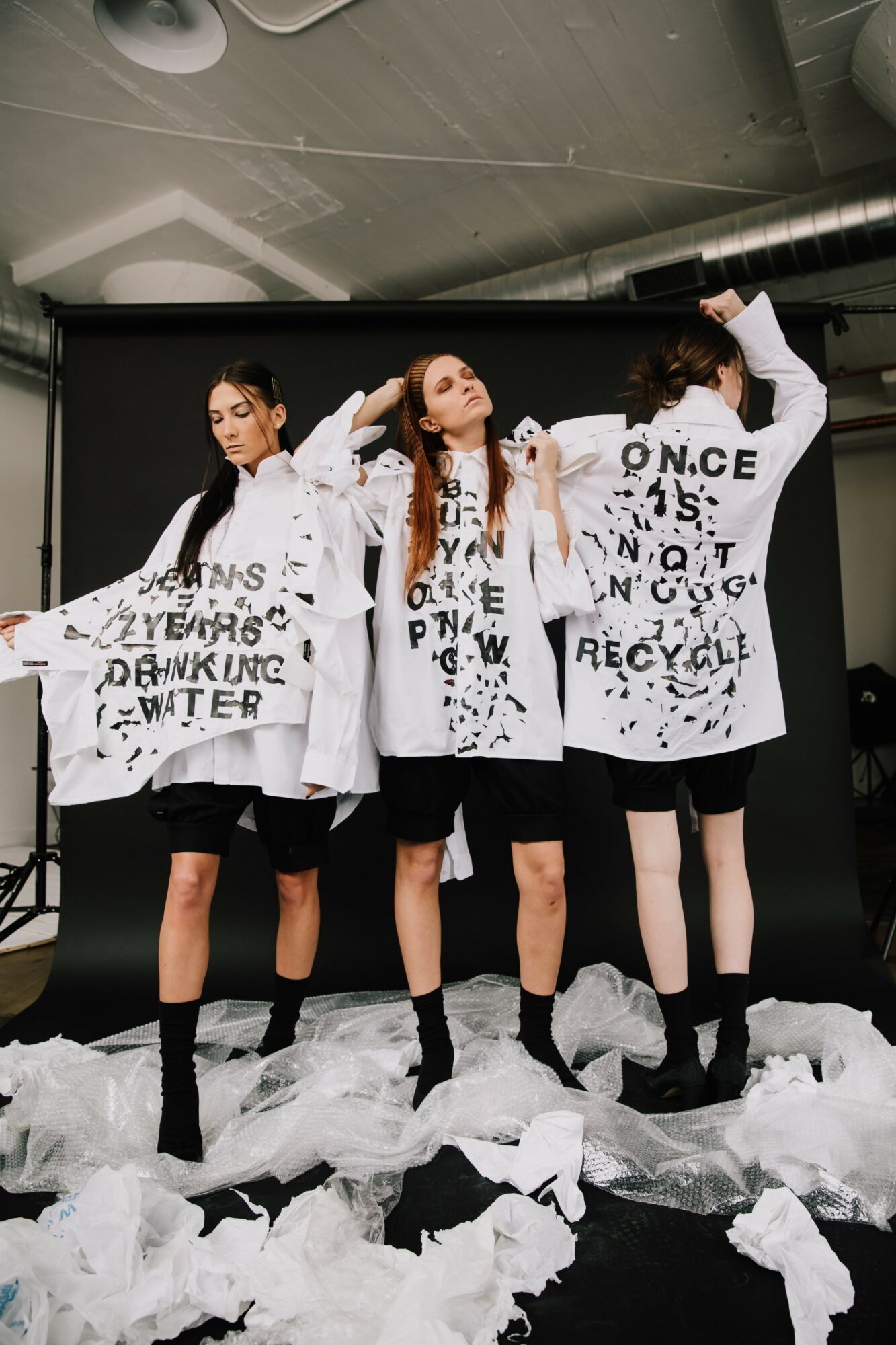 Four people wear oversized white jackets with black text, standing against a black background, with crumpled paper on the floor.