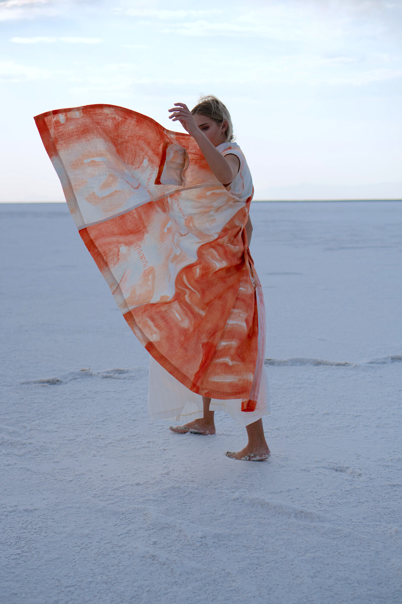 Person standing on salt flat holding a large orange and white patterned fabric, with a clear sky and horizon in the background.