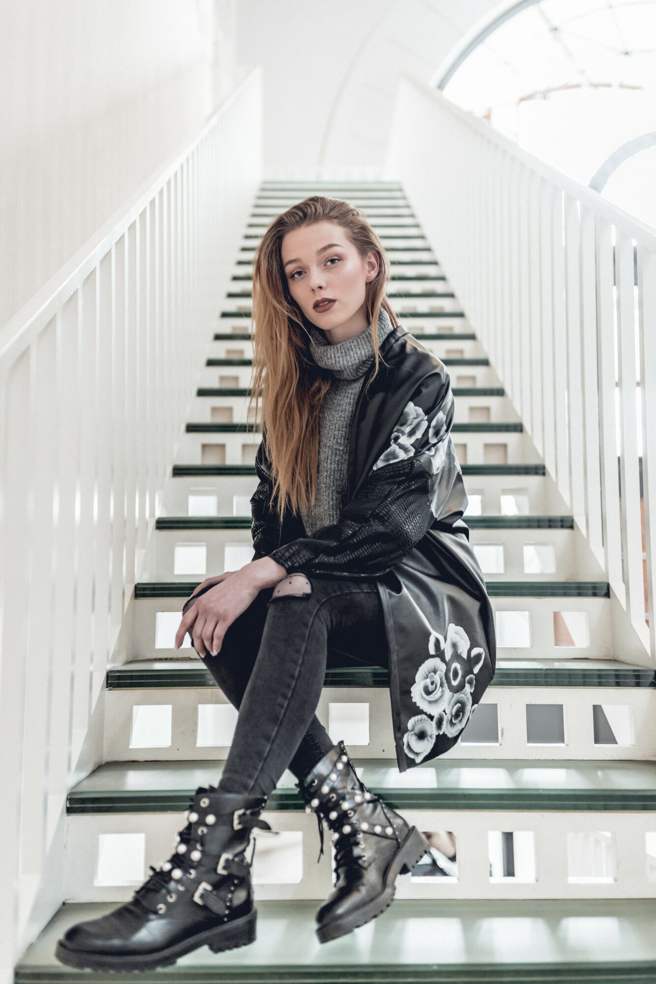 Young woman with long hair sitting on stairs, wearing a black and gray jacket, black pants, and combat boots, inside a modern building.