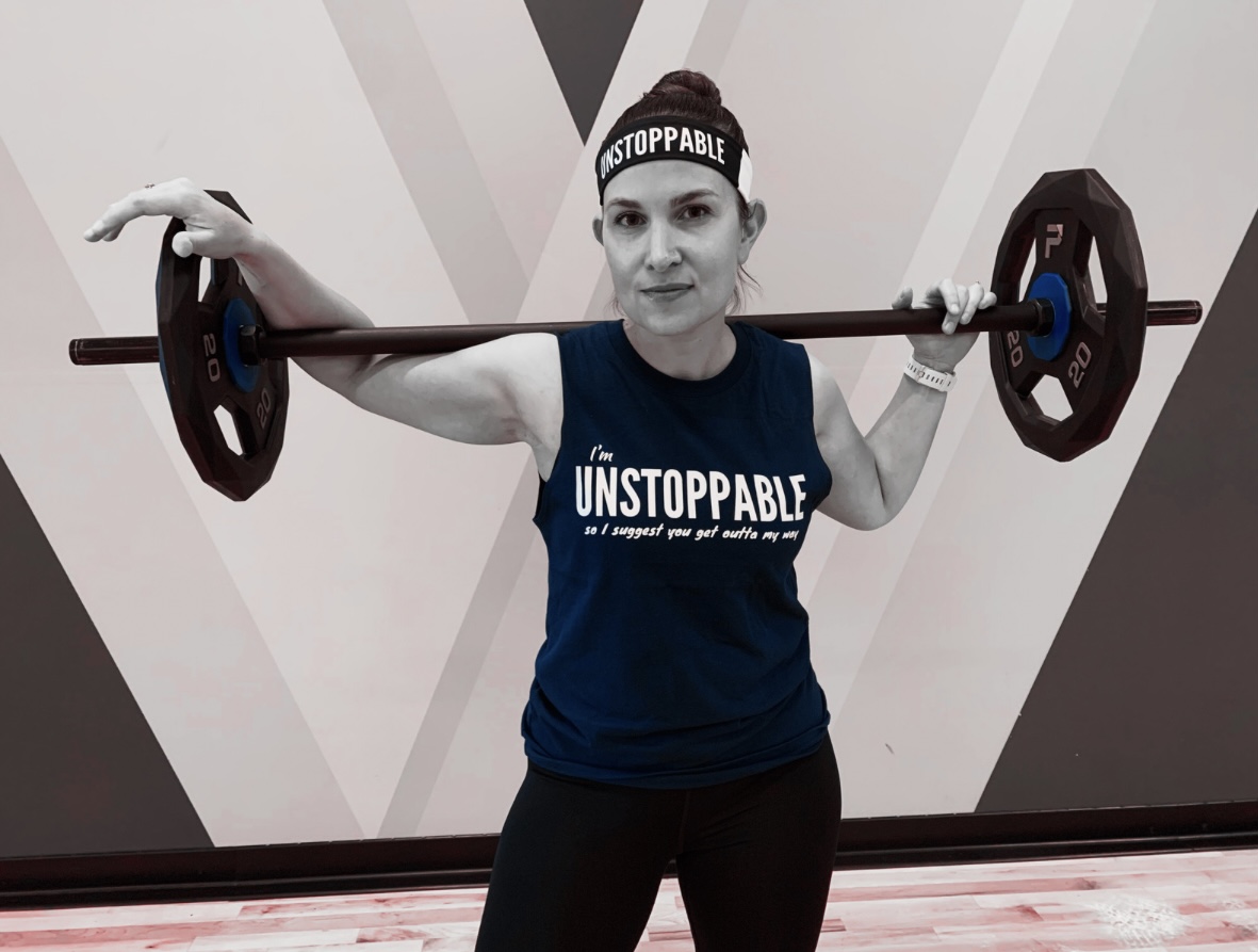 Woman holding a barbell across shoulders, wearing a headband and a sleeveless shirt, in a gym setting.