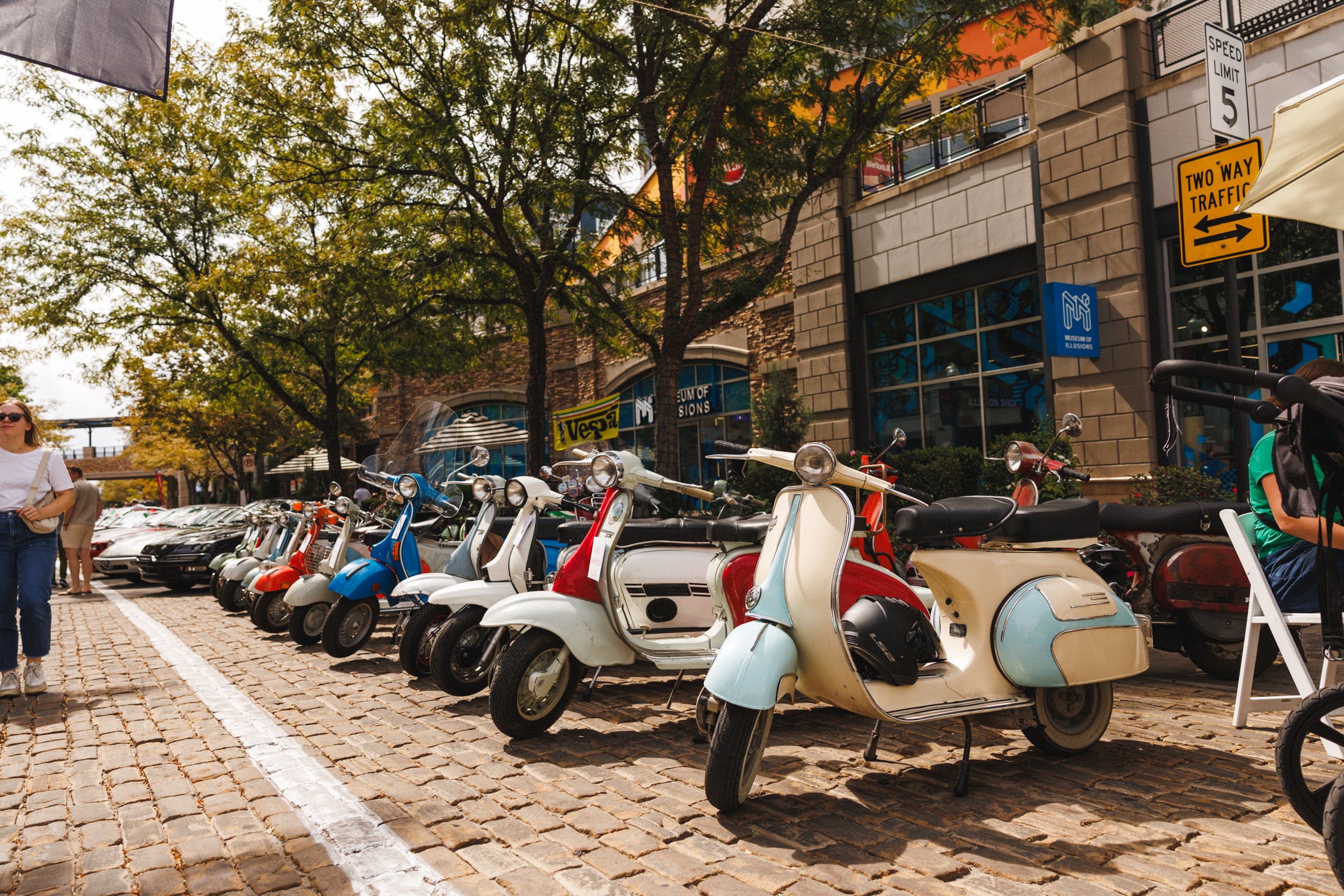 Row of parked scooters along a sidewalk with trees and buildings in the background.