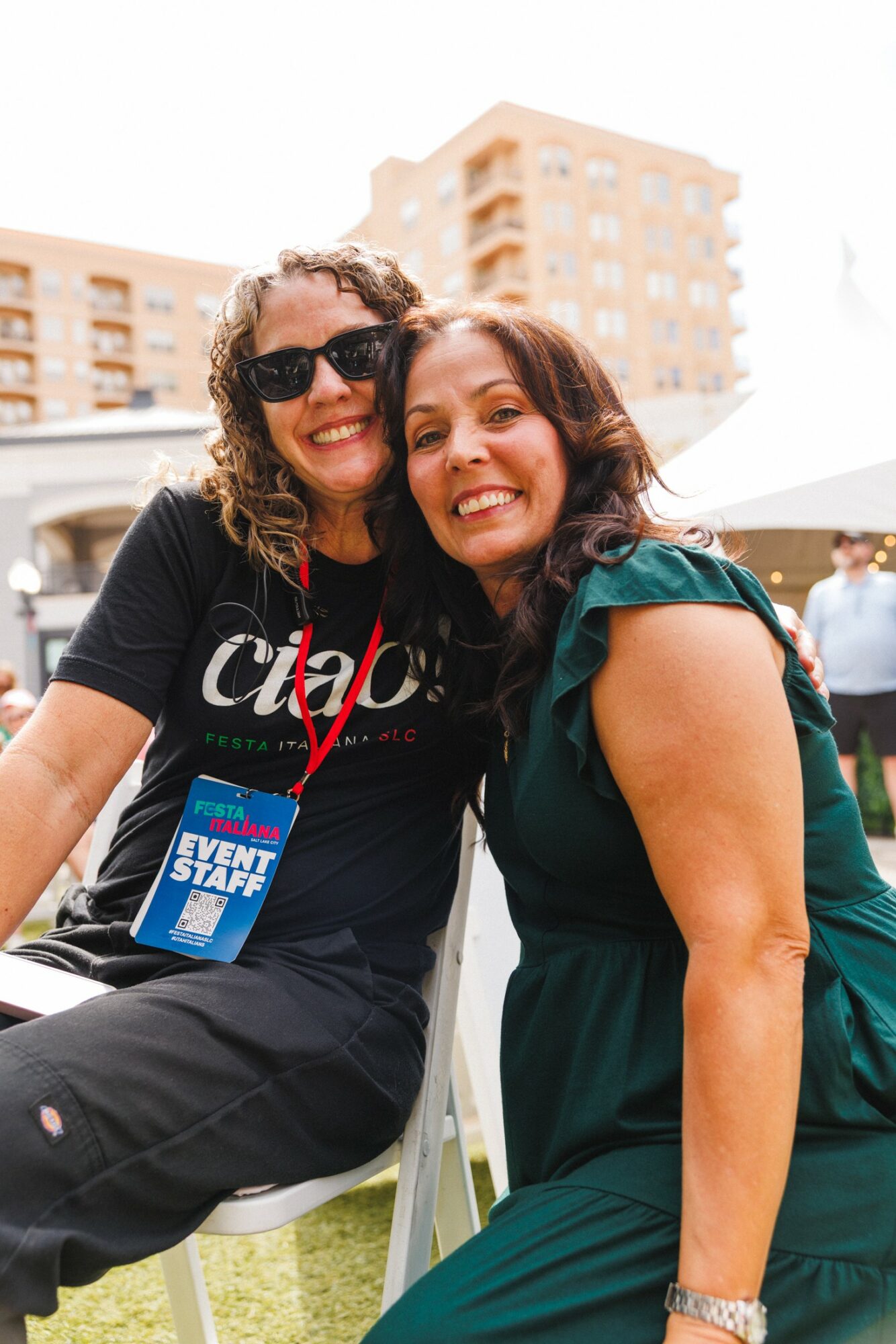 Two women smiling outdoors, one wearing sunglasses and a black shirt, the other in a green dress, with buildings in background.