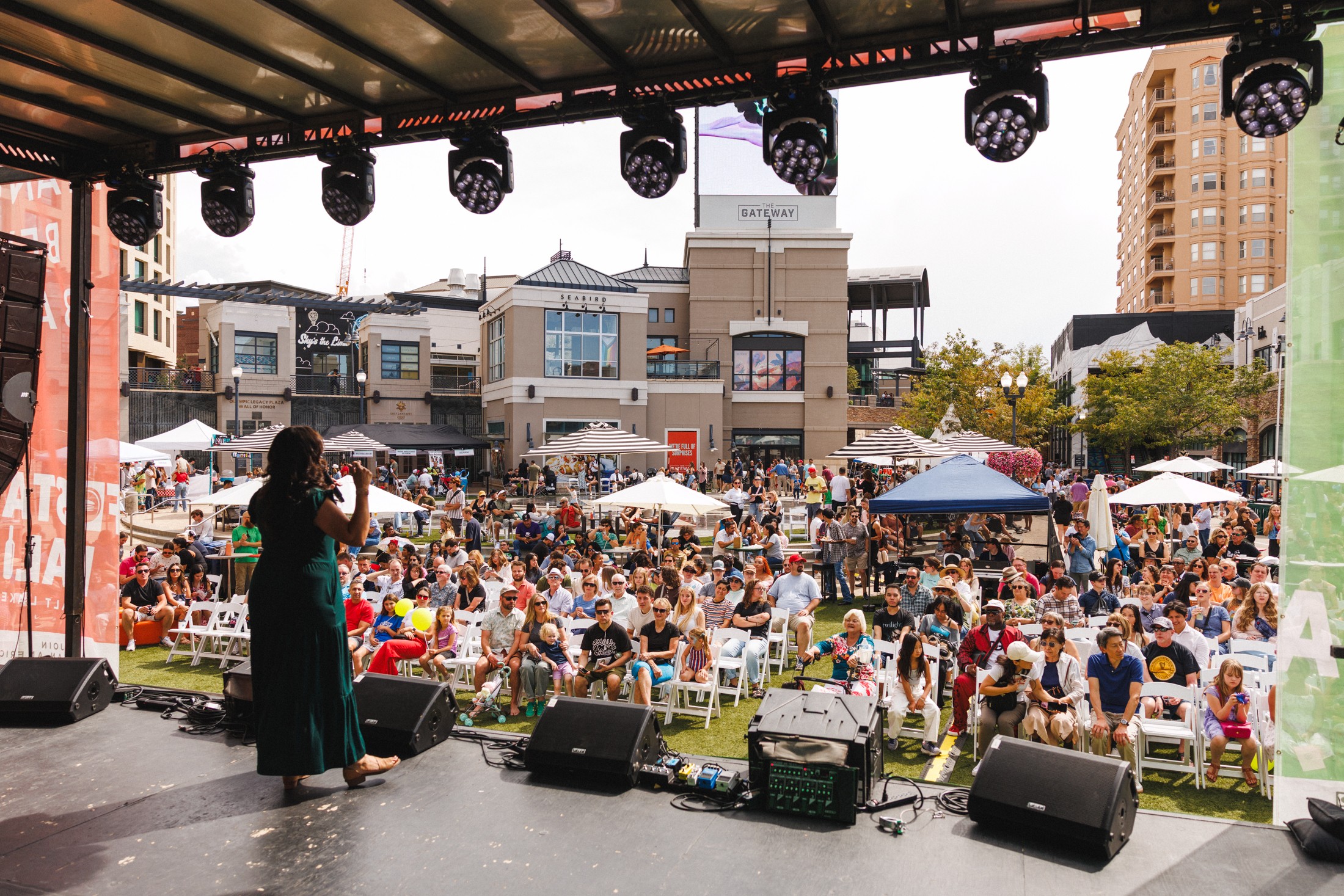 Performer on stage facing a large outdoor crowd with tents and buildings in background.