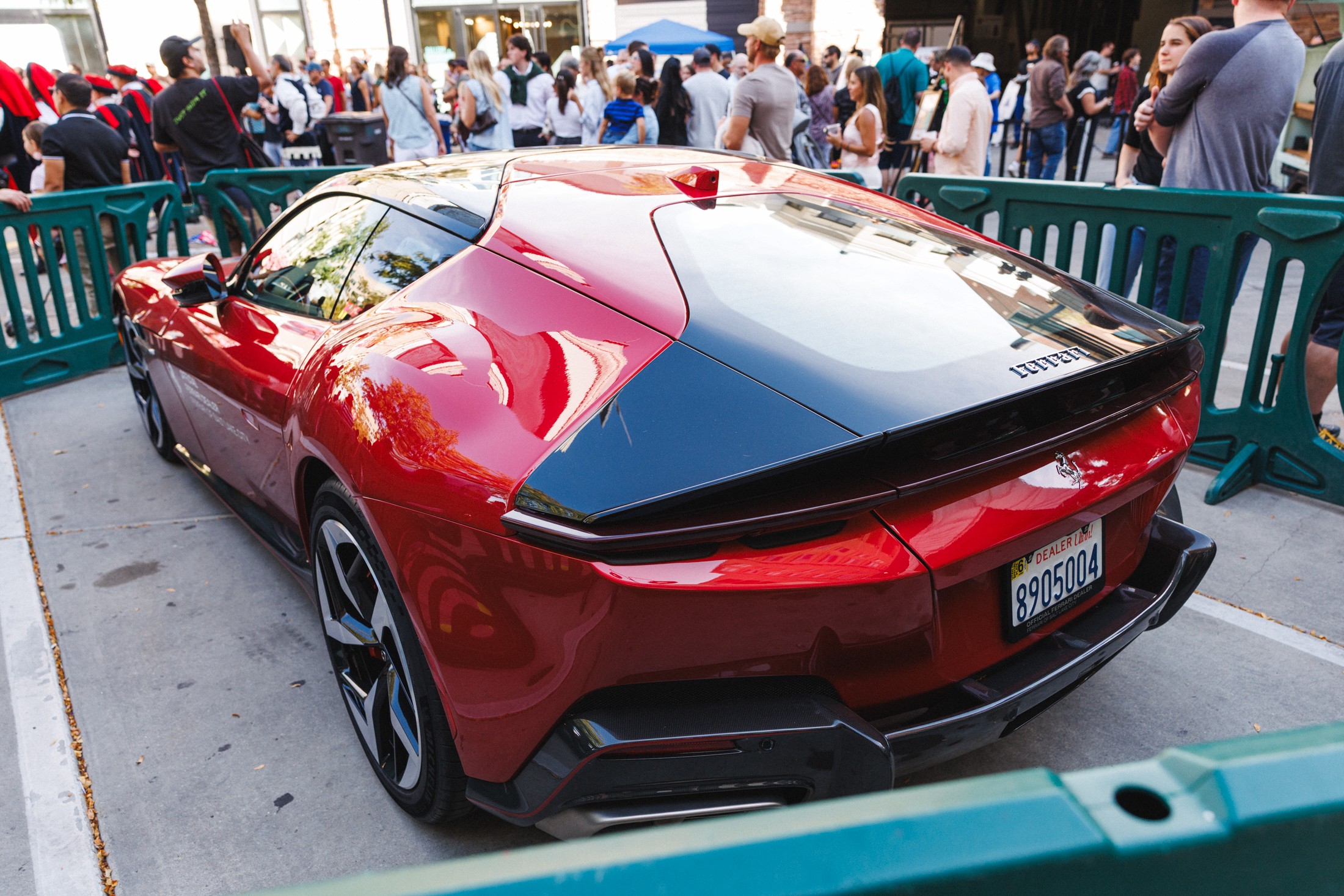 Red sports car parked outdoors with a crowd in the background, surrounded by green barriers.