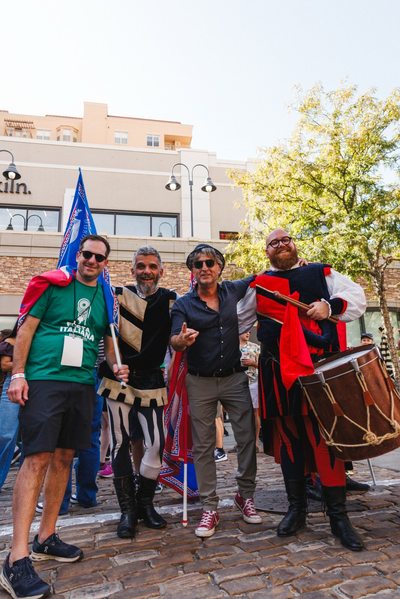 Group of four people standing outdoors, one holding a flag, with buildings and trees in background.