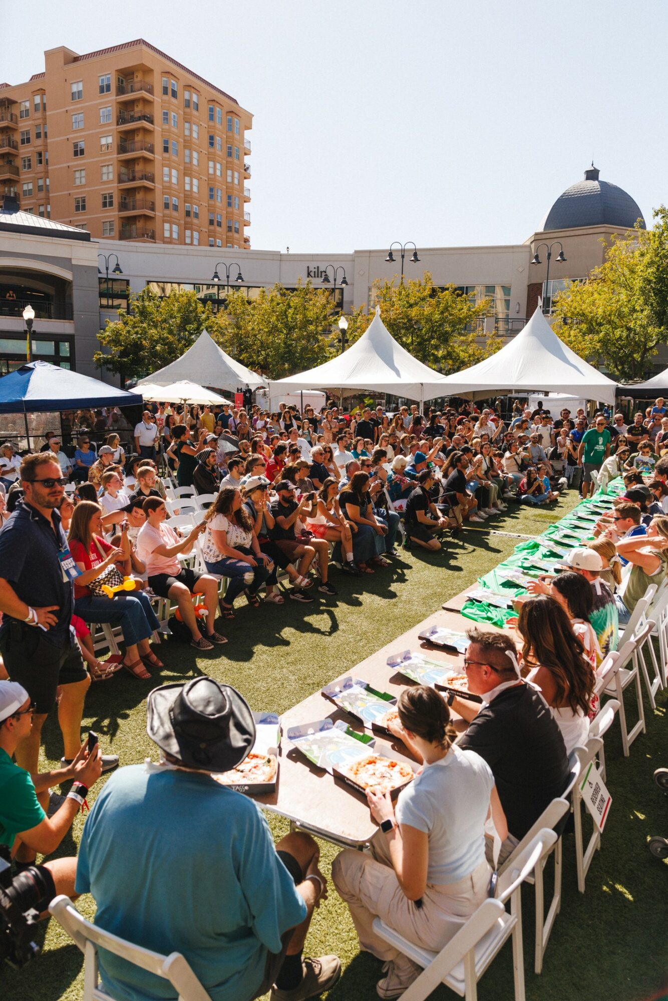 Crowd of people seated outdoors at tables with tents, in an urban area with buildings and trees, sunny day.