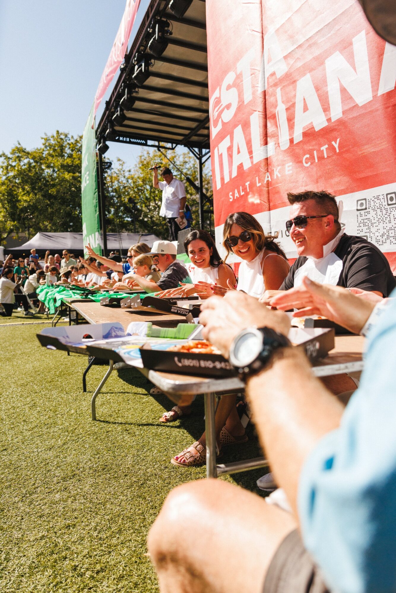 People sitting at a long table outdoors during daytime event, with a large red banner in the background, some wearing sunglasses.