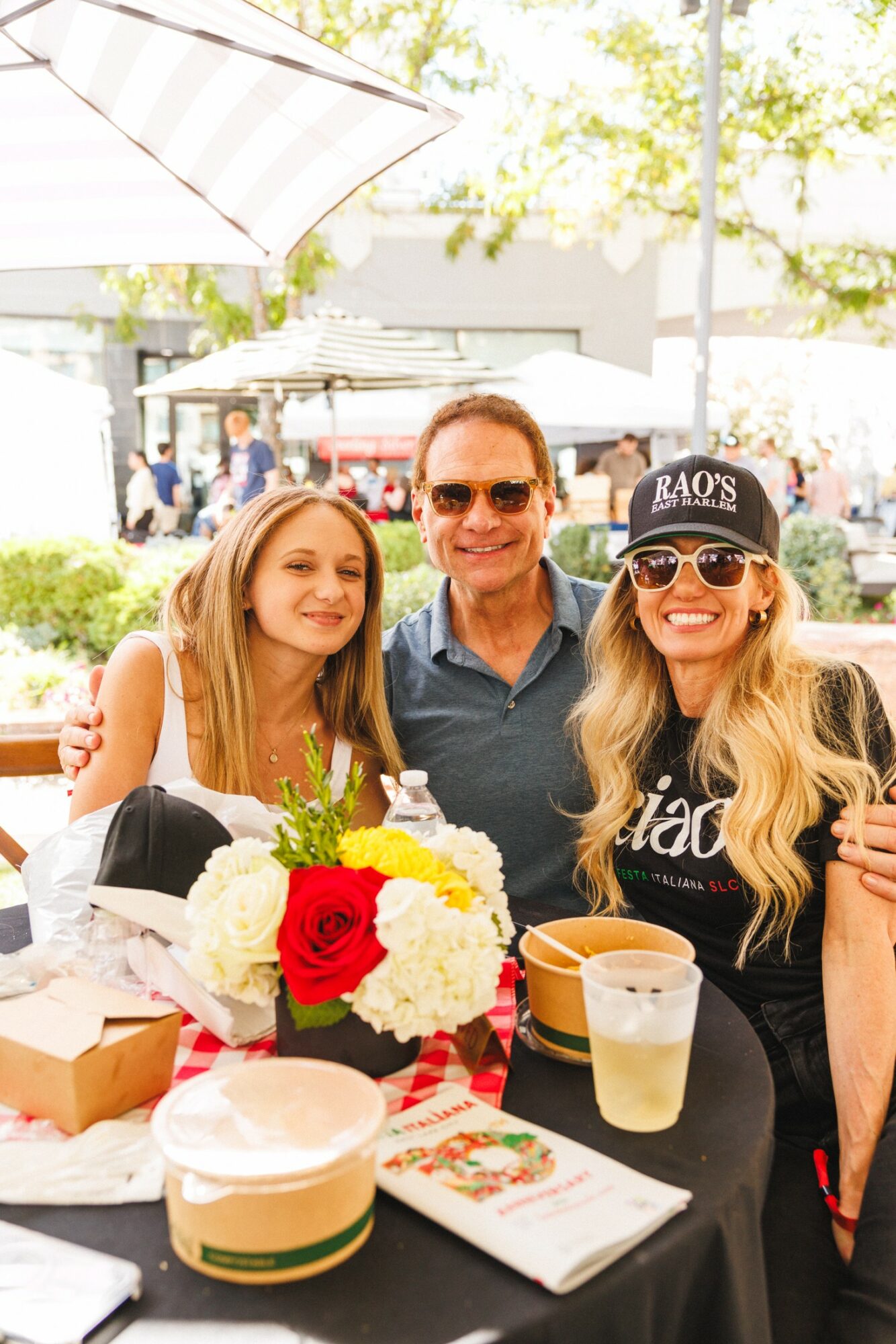 Three people sitting at a table outdoors with flowers, drinks, and a hat, smiling in sunlight.