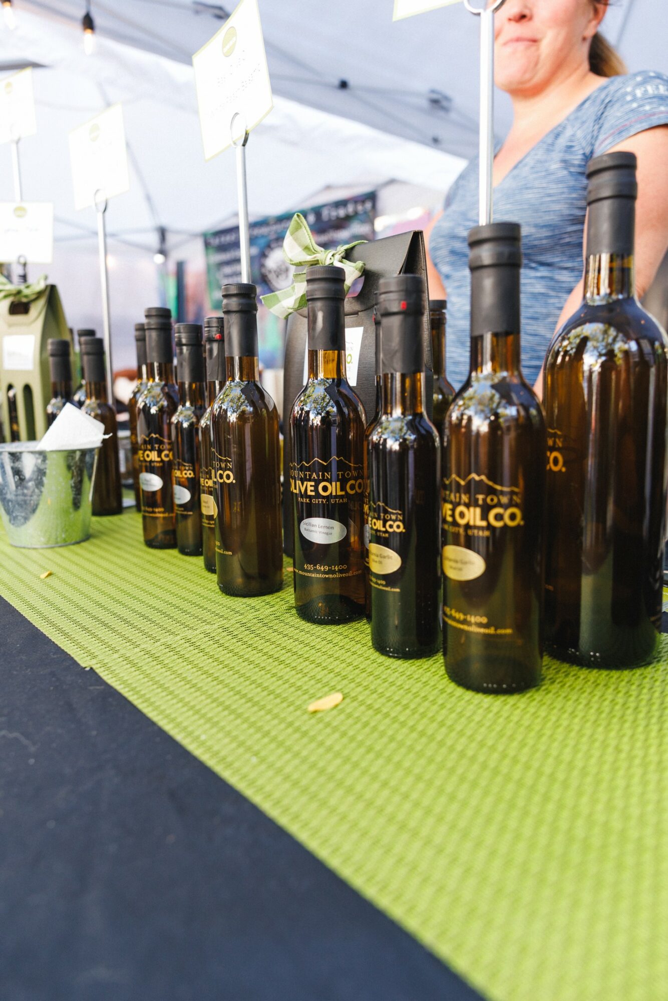Row of olive oil bottles on a green tablecloth at an outdoor market stall, with a person partially visible in the background.