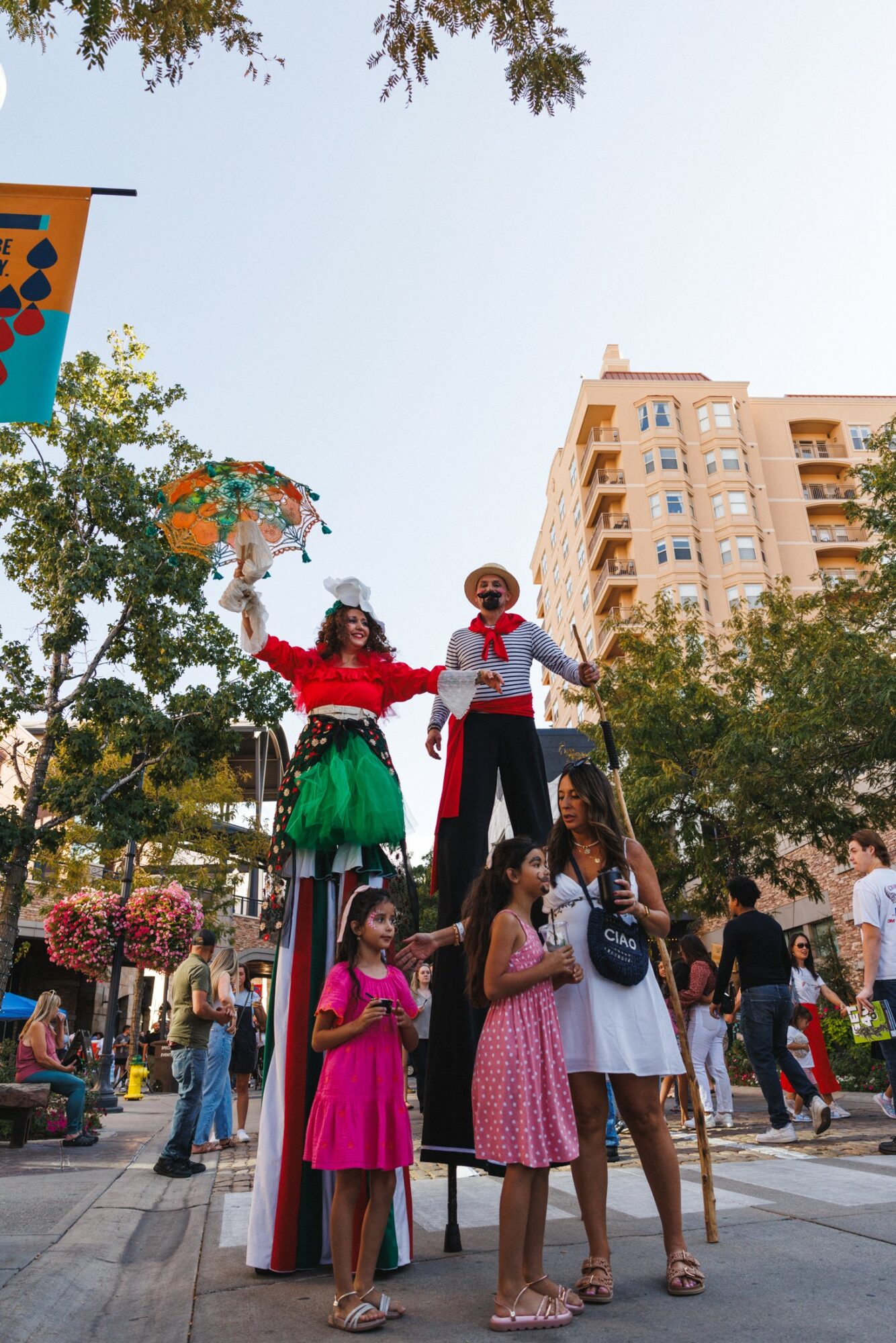 Two large puppets with children and adults in a city square, trees, and a tall building in the background.