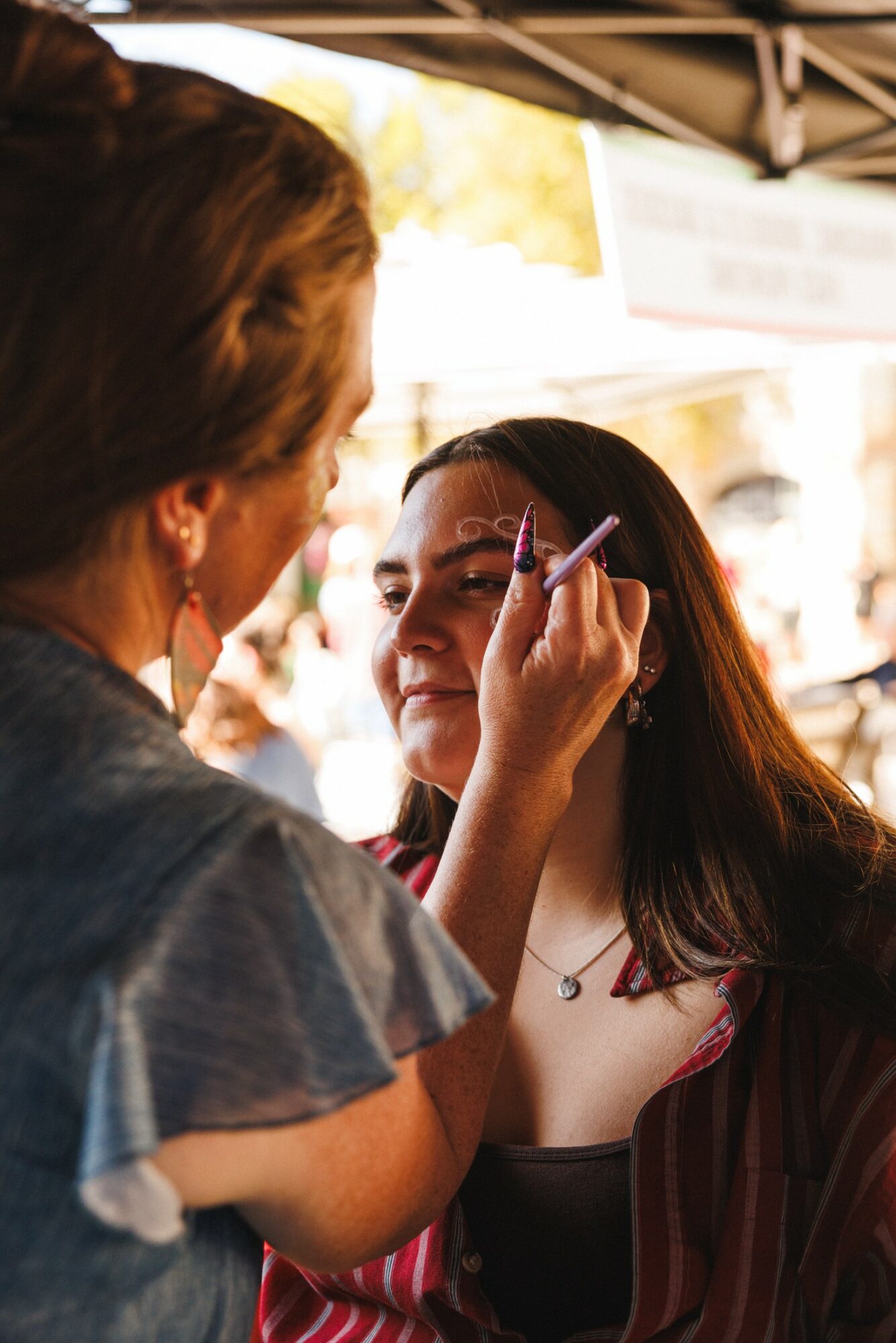 Person applying makeup to a woman at an outdoor event, with a tent and blurred background behind them.