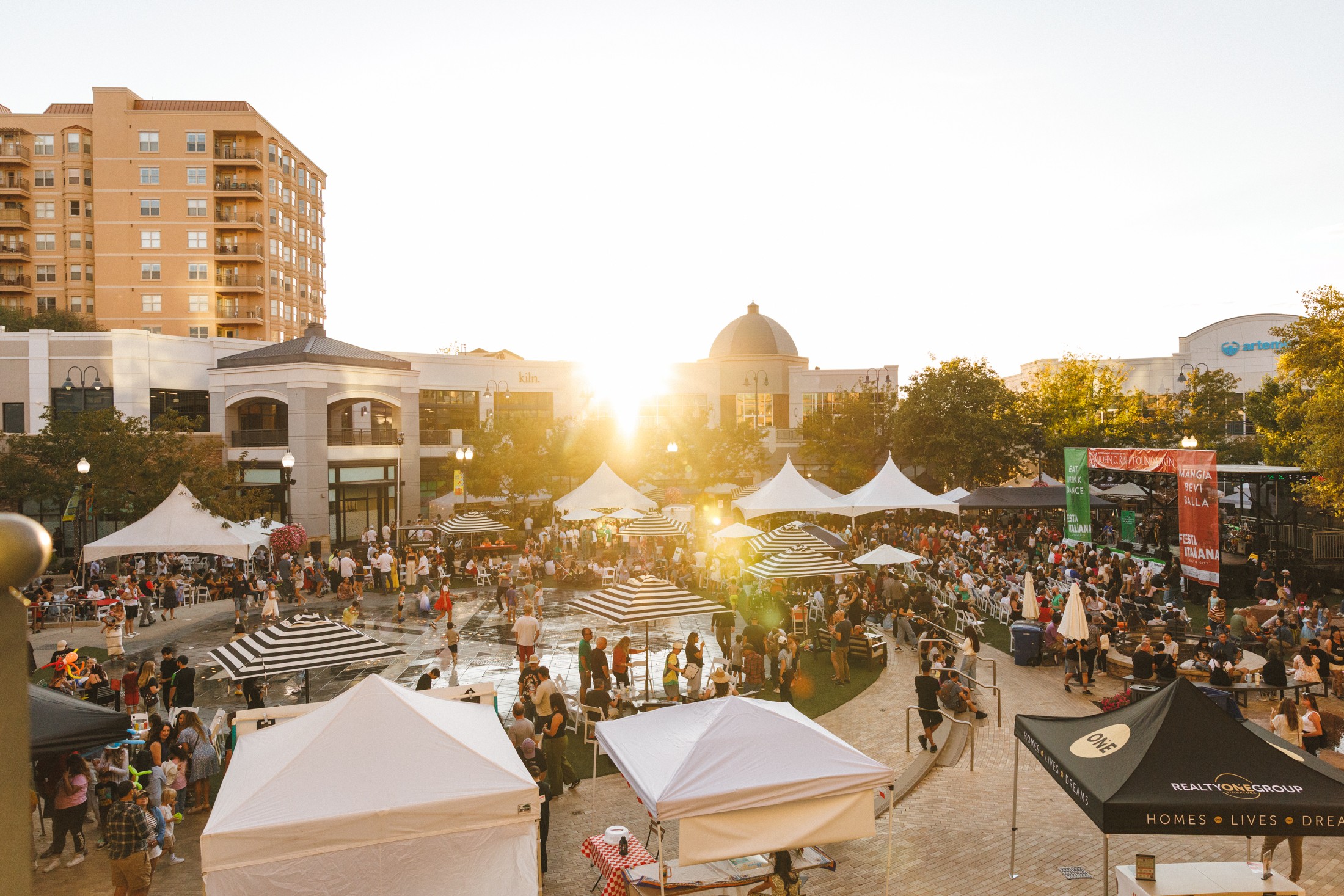 Crowd gathered at outdoor event with tents, buildings, and trees, sunlight shining in the background.