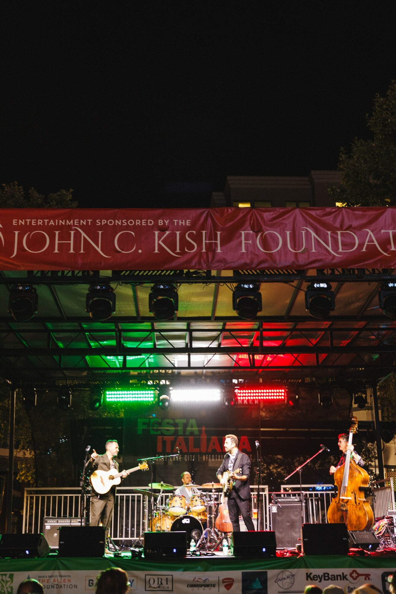 Band performs on stage with colorful lights, audience in front, night sky above, trees in background.