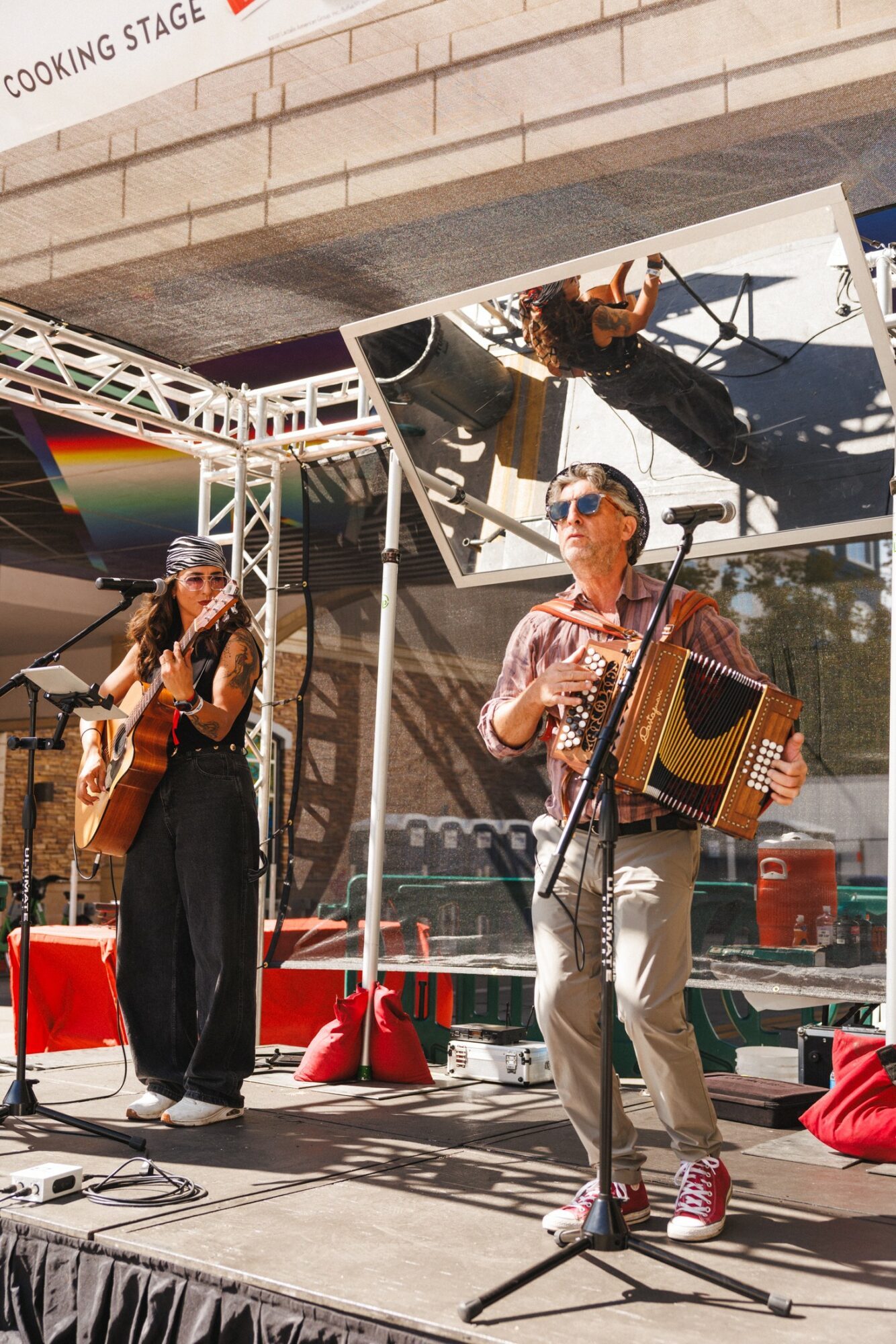 Two musicians perform on an outdoor stage, one playing guitar and the other playing an accordion, with a reflective ceiling above.
