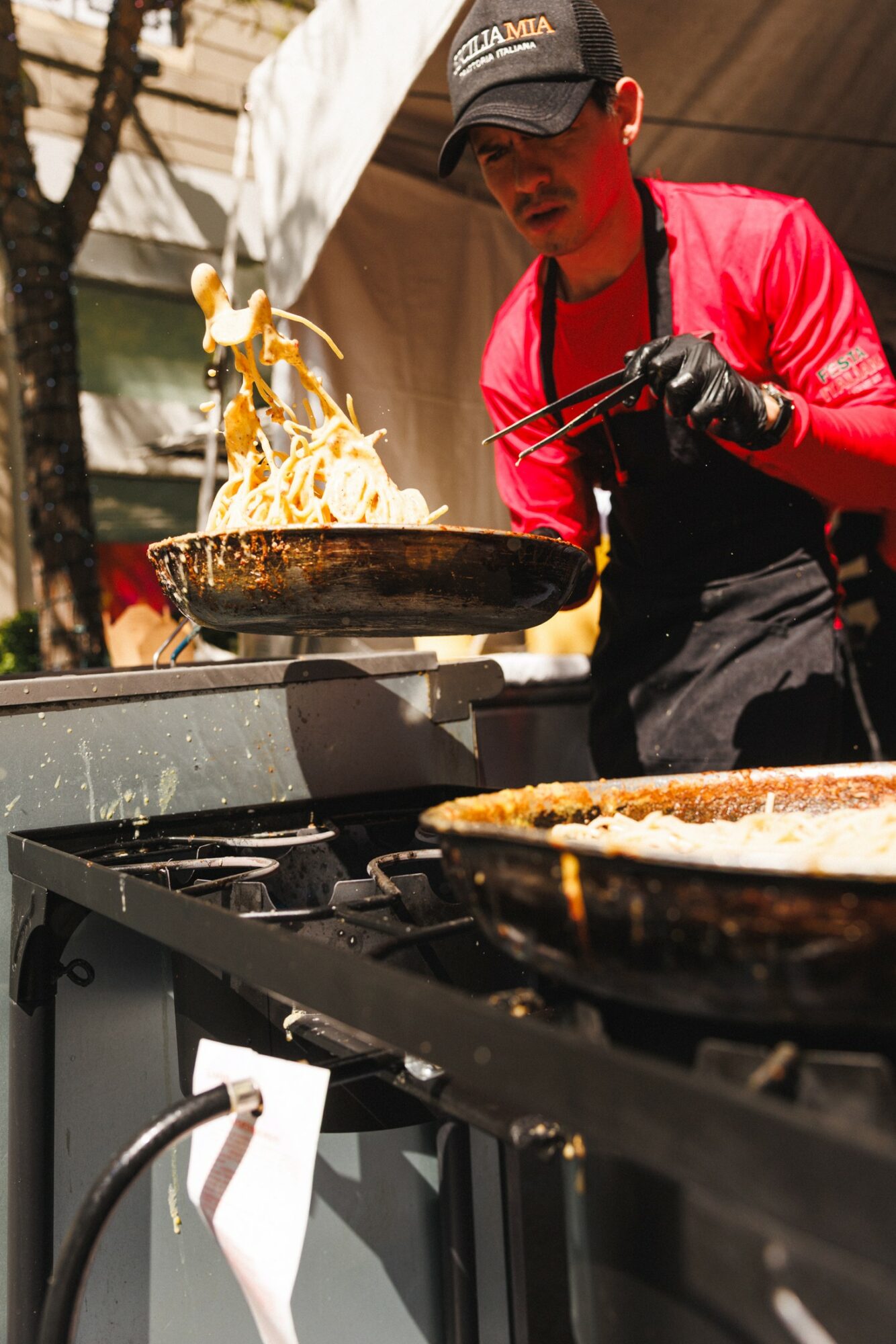 Chef in red shirt and black apron flips food in a pan outdoors, flames visible, with another pan nearby.