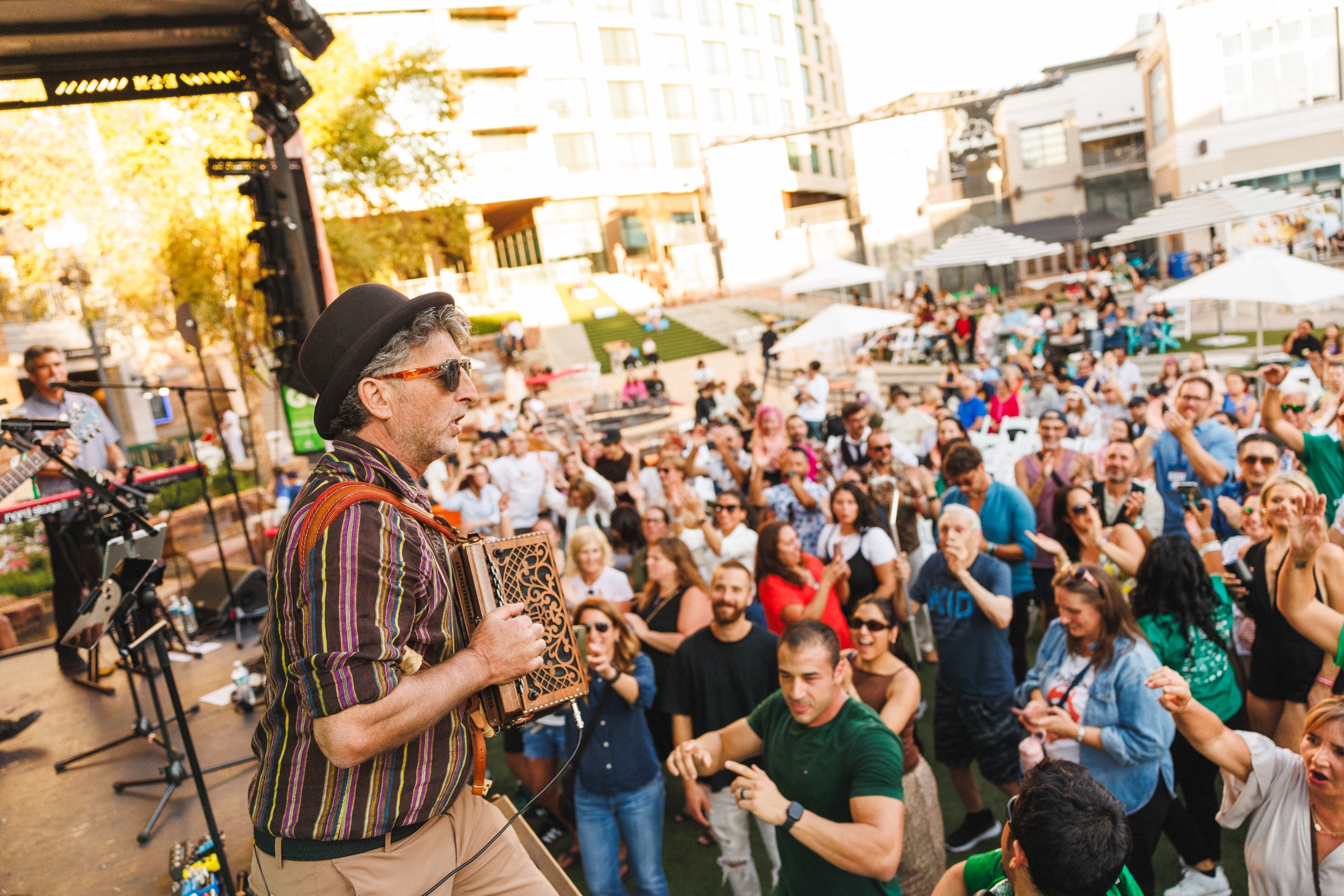 Performer with a hat and striped shirt playing an instrument on stage, crowd dancing outdoors in a lively urban setting.