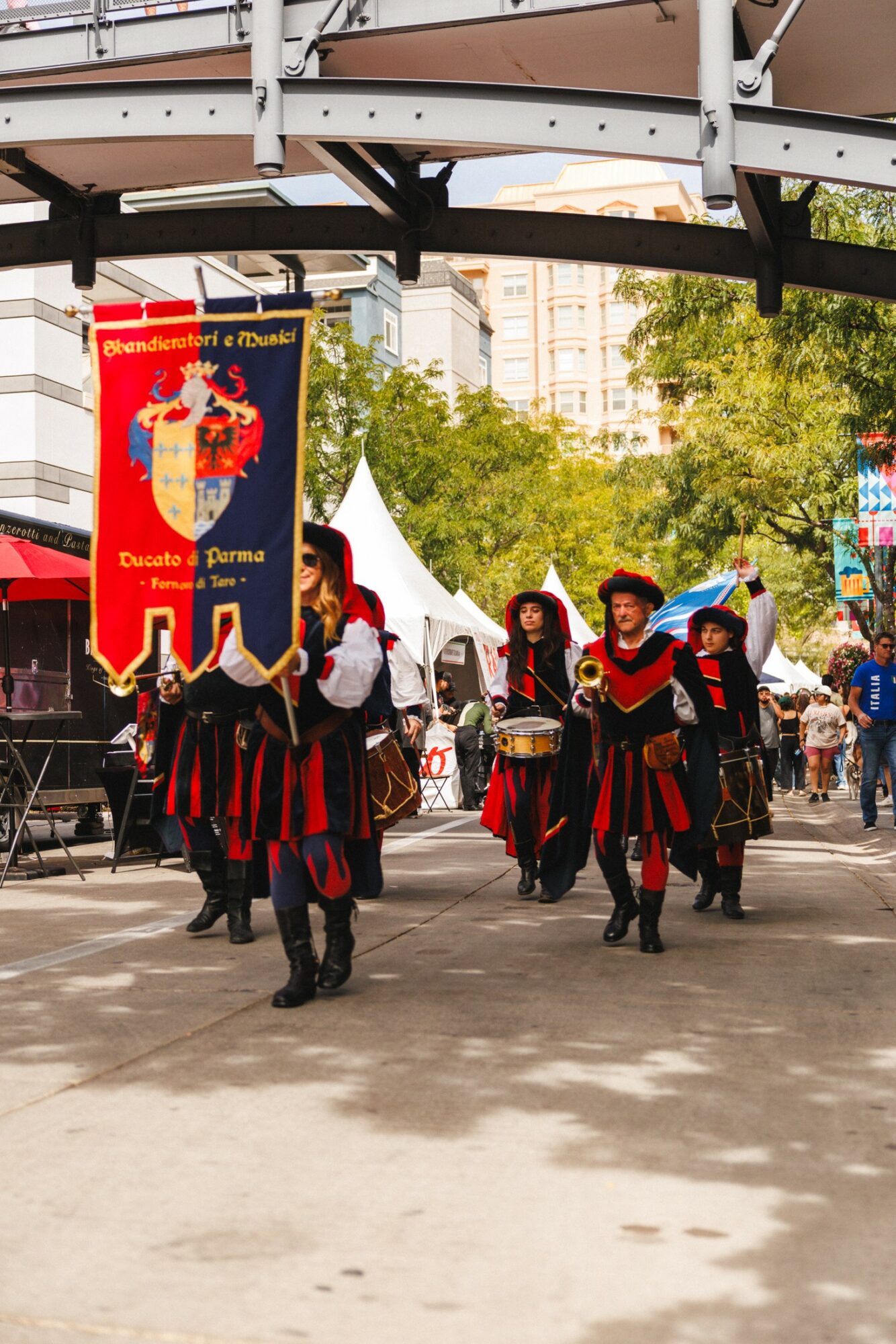 Group of people in historical costumes walking outdoors under a bridge, with tents and trees in the background.