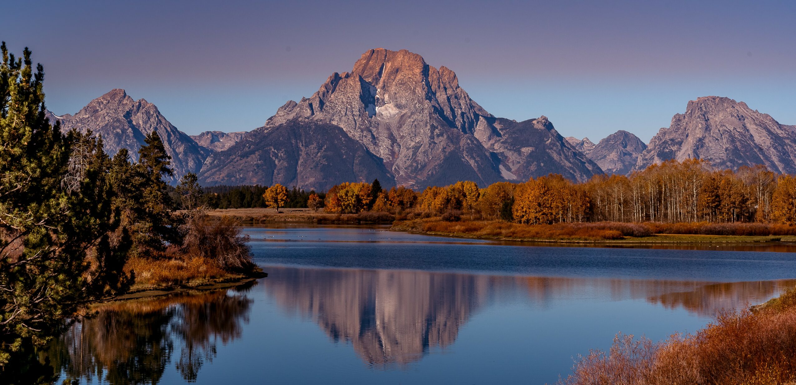 Mountain range with peaks reflected in a calm river, surrounded by trees and clear sky.