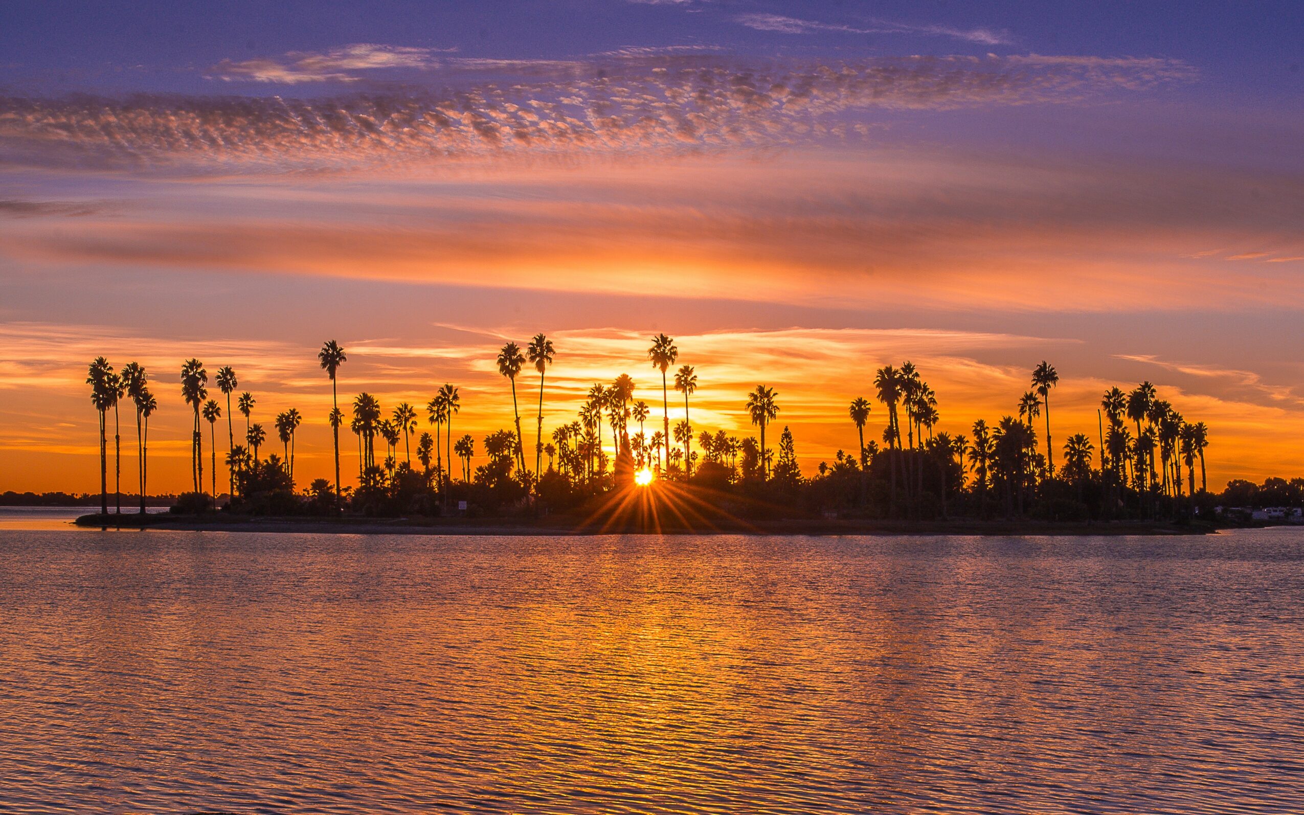 Sunset over water with silhouettes of palm trees on the horizon, colorful sky with orange, pink, and purple hues.