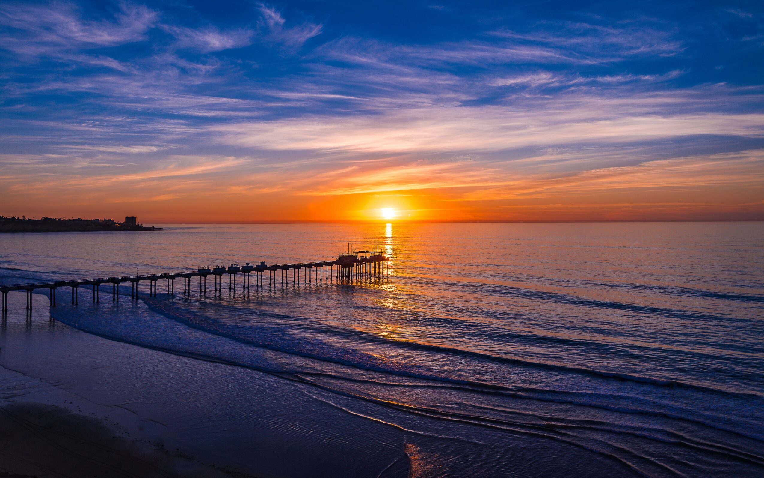 Sunset over calm ocean with a pier extending into the water, colorful sky with clouds, and gentle waves on the beach.