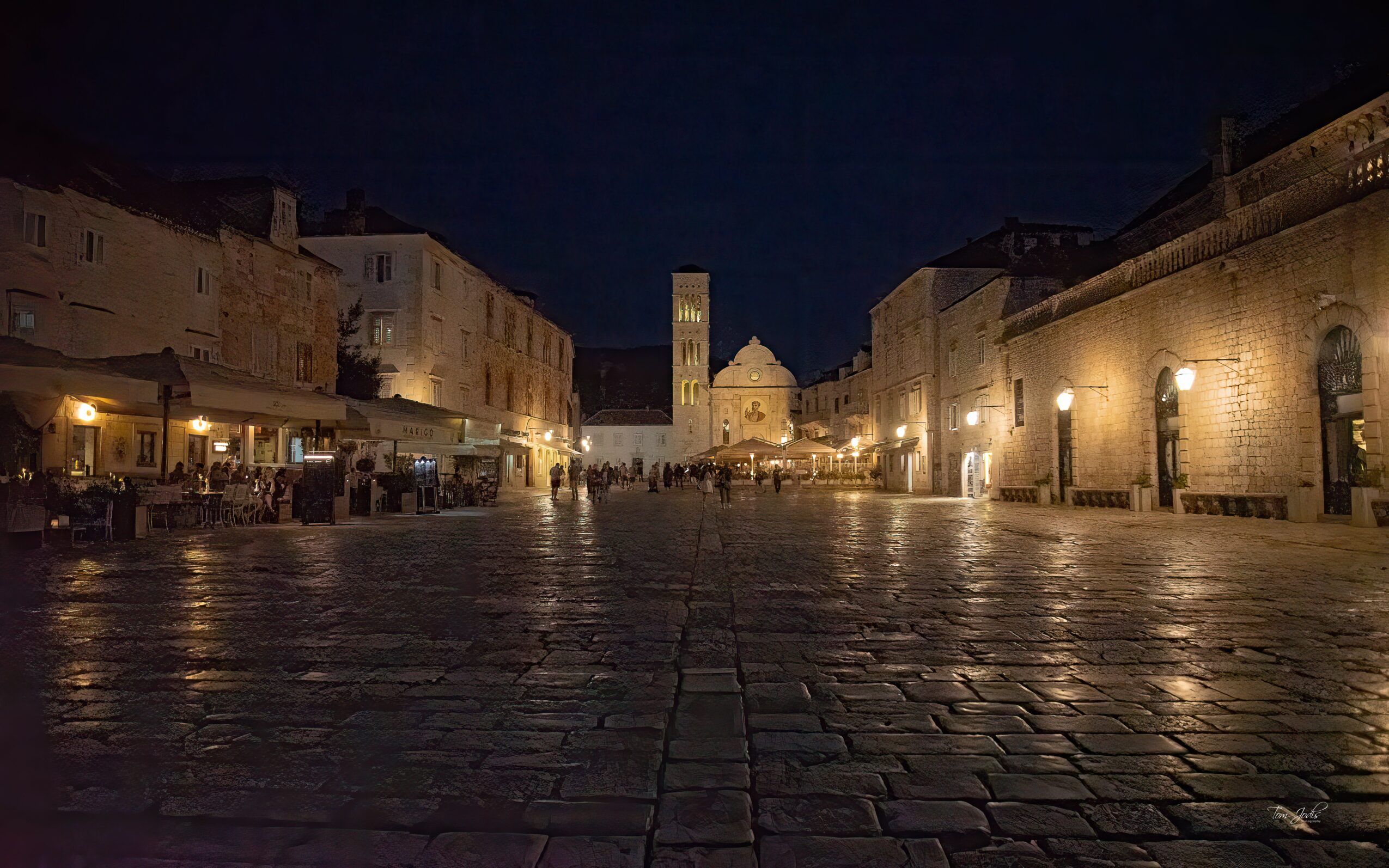 Night scene of a historic town square with cobblestone pavement, illuminated buildings, and a tower in the distance.