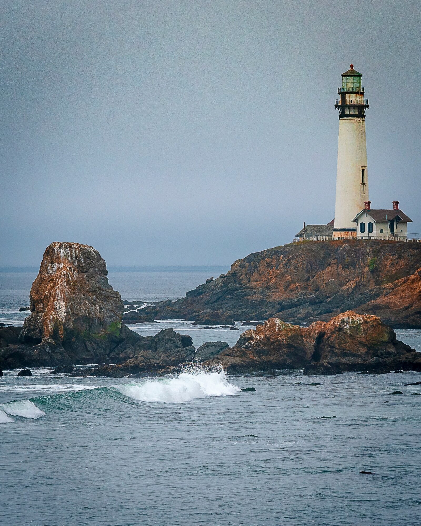Lighthouse on rocky coastline with ocean waves, cloudy sky, and rugged terrain.