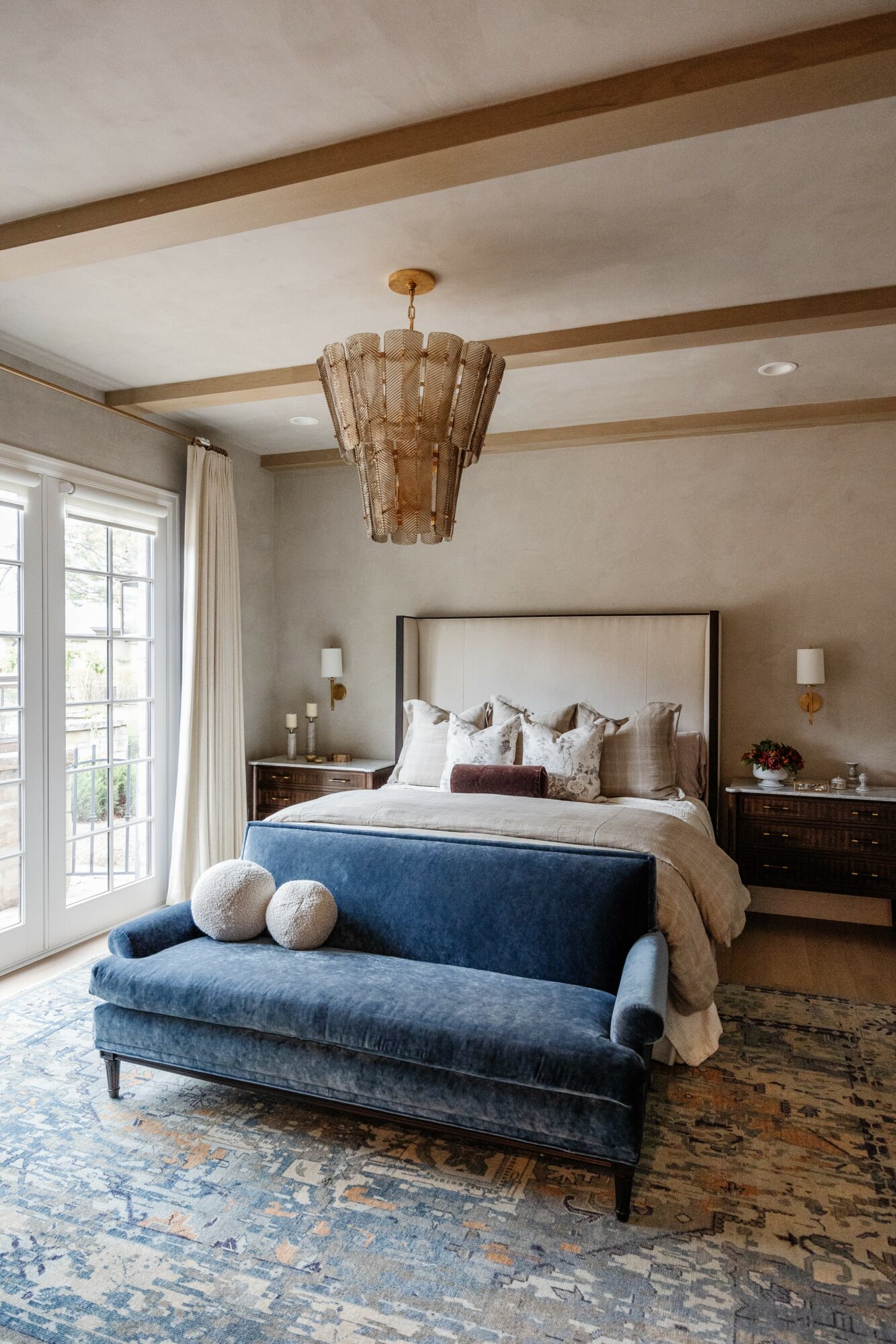 Bedroom with bed, blue bench, chandelier, and large window, neutral walls, and wooden flooring.