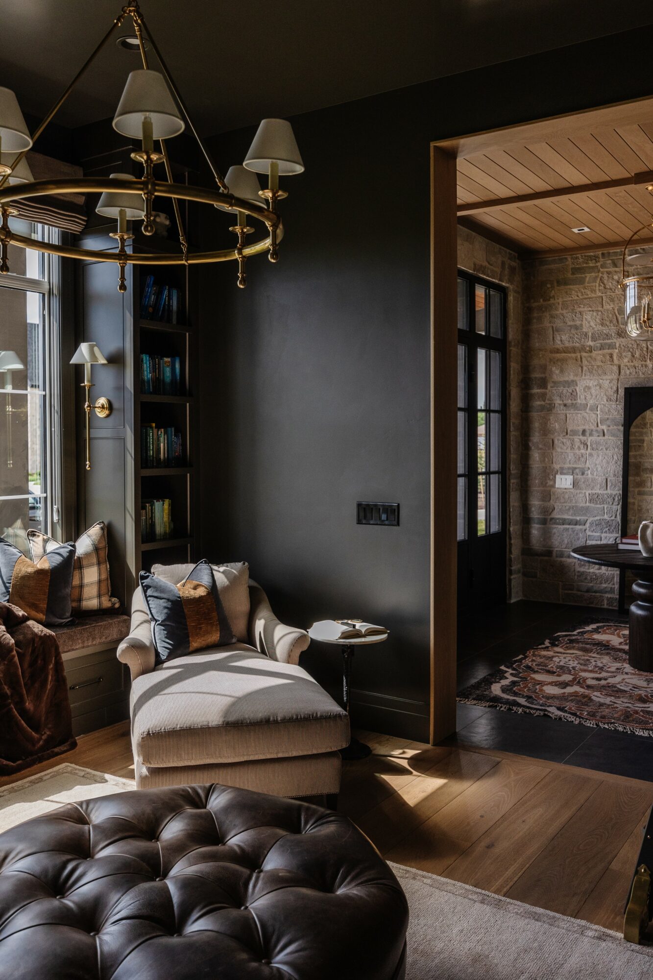 Living room with dark wall, white armchair, window with curtains, and chandelier, adjacent to a room with brick wall and wooden ceiling.