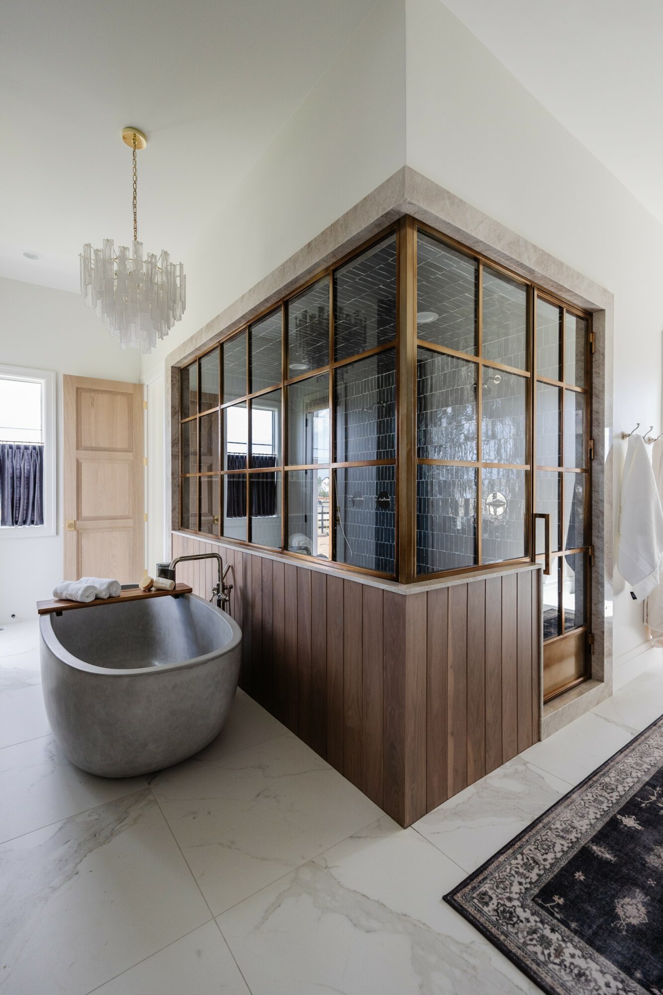 Corner of a room with a wooden and glass partition, a freestanding bathtub, a chandelier, and a rug.