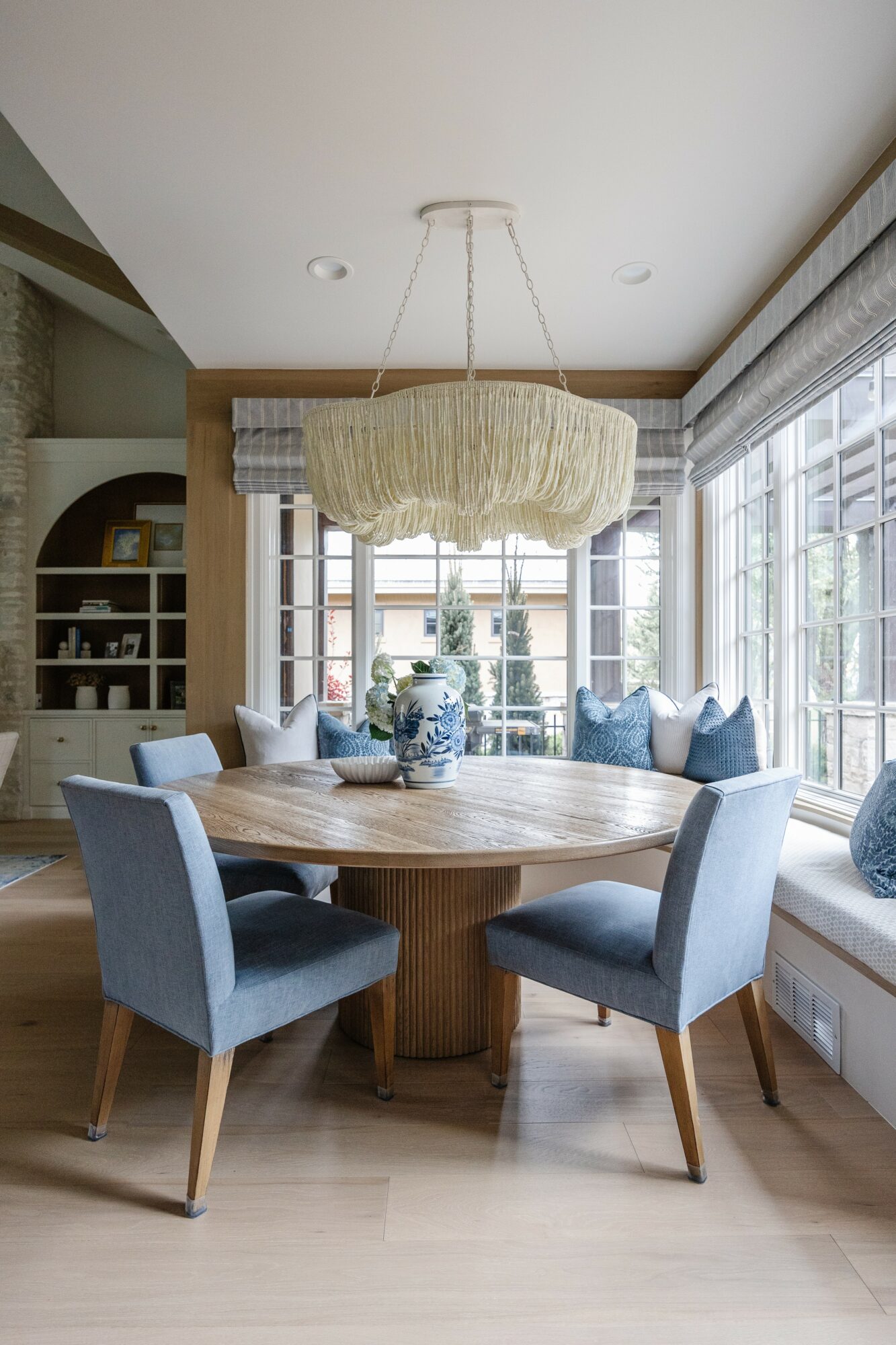 Round wooden dining table with five blue upholstered chairs in a bright room with large windows and a window seat with cushions.