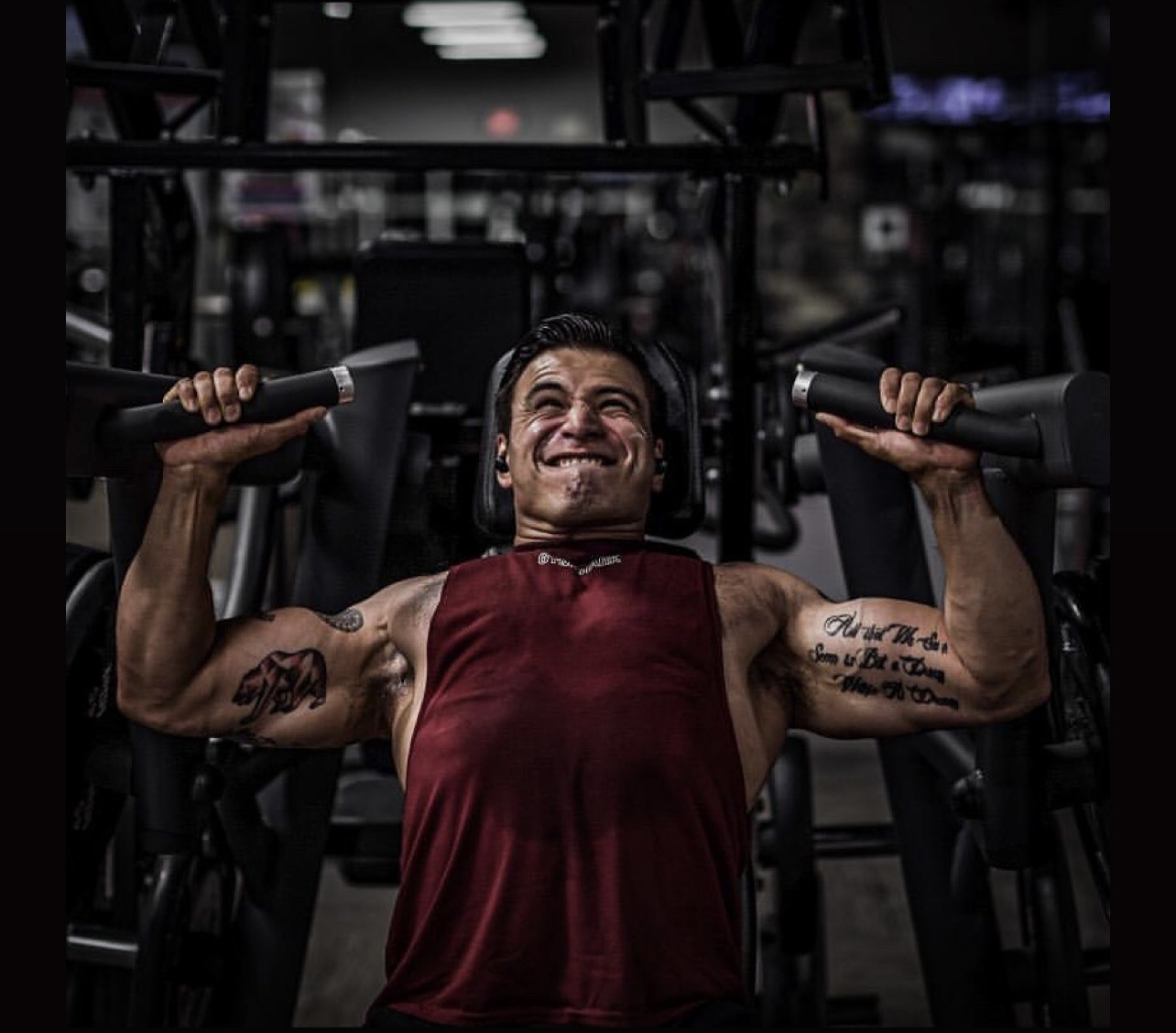Person lifting weights in gym, smiling, wearing a sleeveless shirt, with tattoos on arms, dark background.