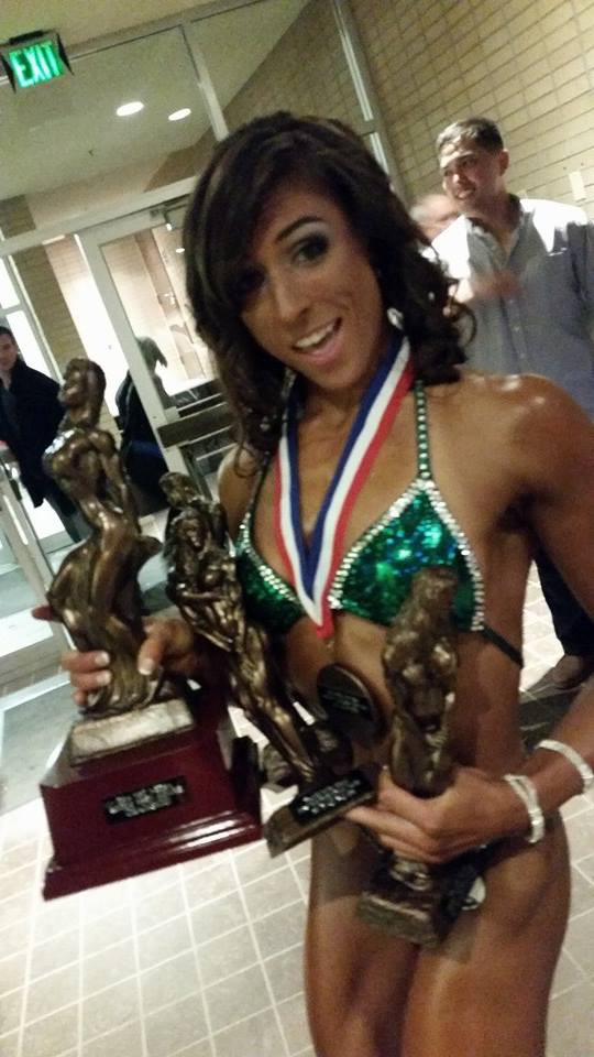 Young woman in a green bikini holding multiple trophies and wearing a medal, smiling at the camera.