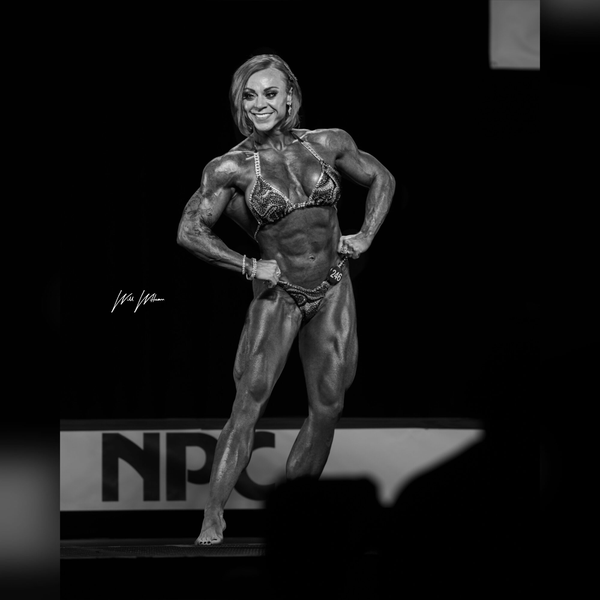 Female bodybuilder on stage, smiling, posing with hands on hips, muscular physique, black background, stage lighting, in competition attire.