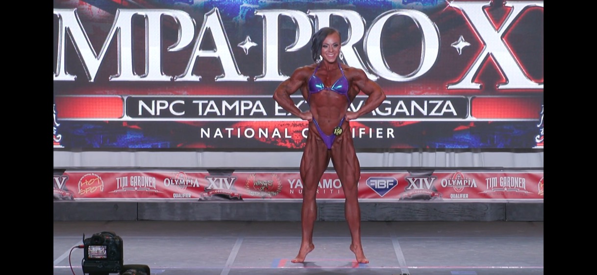 Female bodybuilder on stage in a red bikini, posing in front of a large banner with event text.
