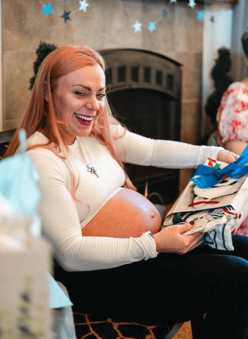 Pregnant woman with red hair opening a gift, smiling, in a decorated room with a fireplace and blue star decorations.
