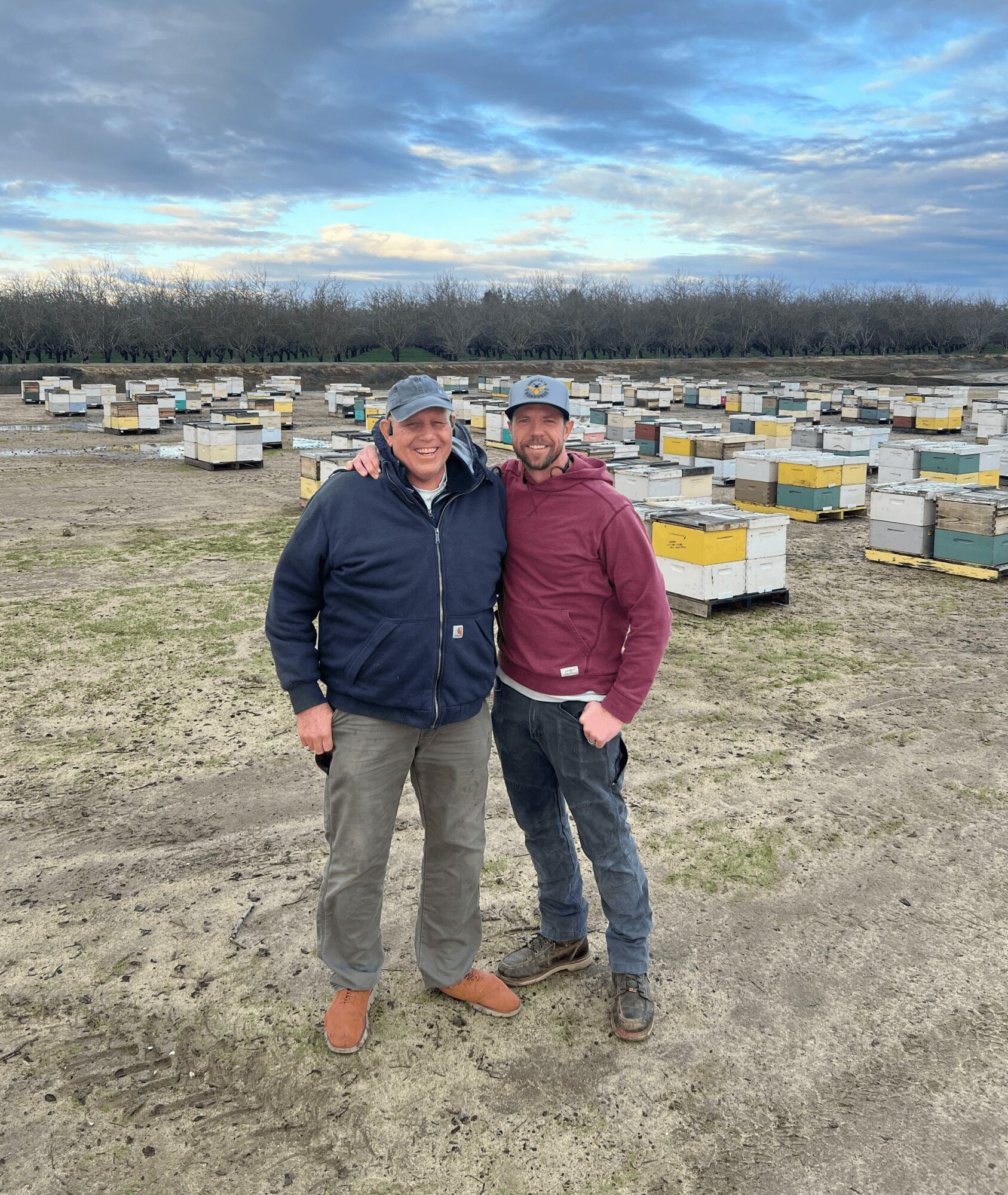Two people stand outdoors in front of numerous beehives, with a cloudy sky and trees in the background.