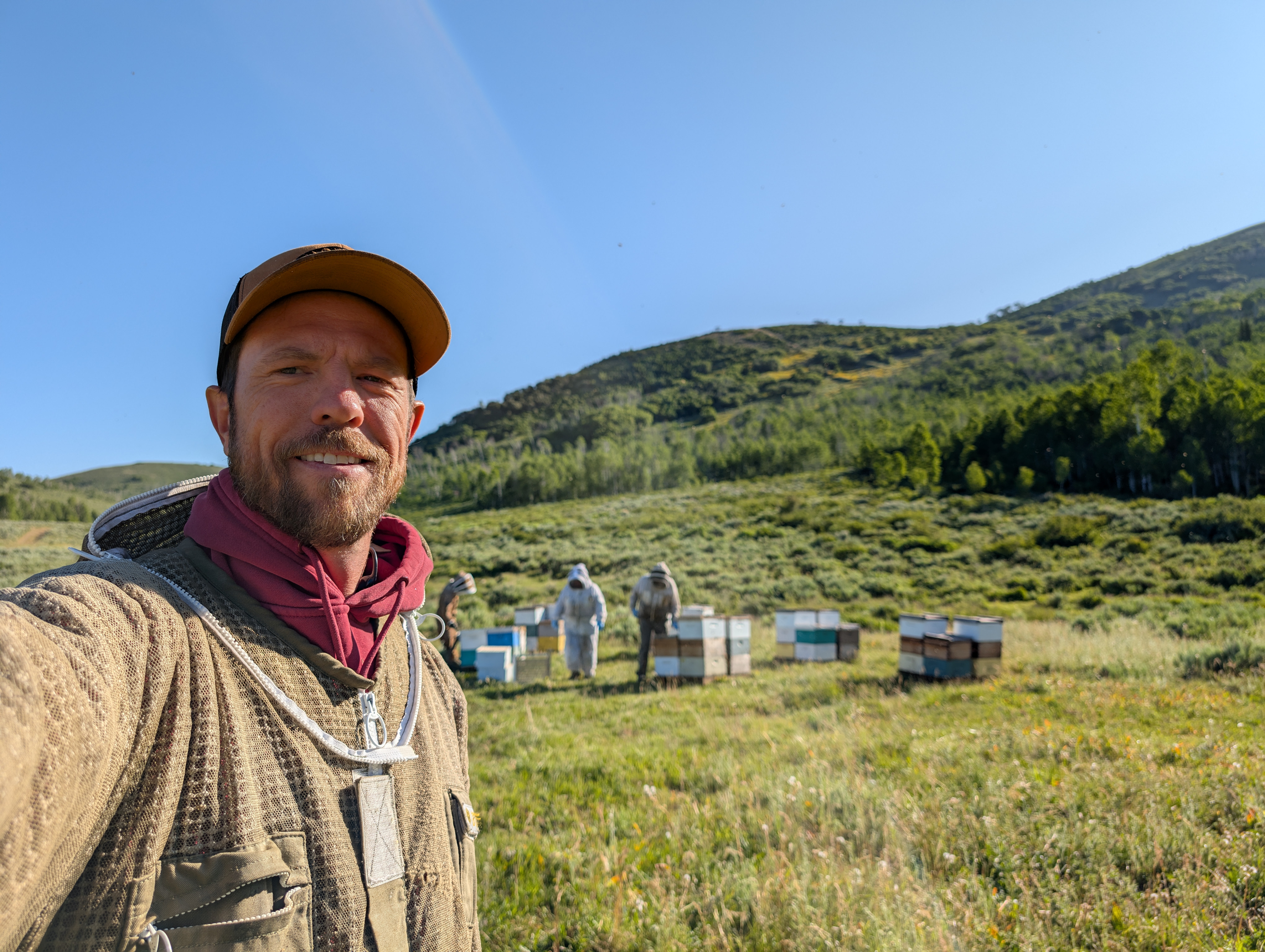 Man taking selfie outdoors with beehives and green hills in background under clear blue sky.