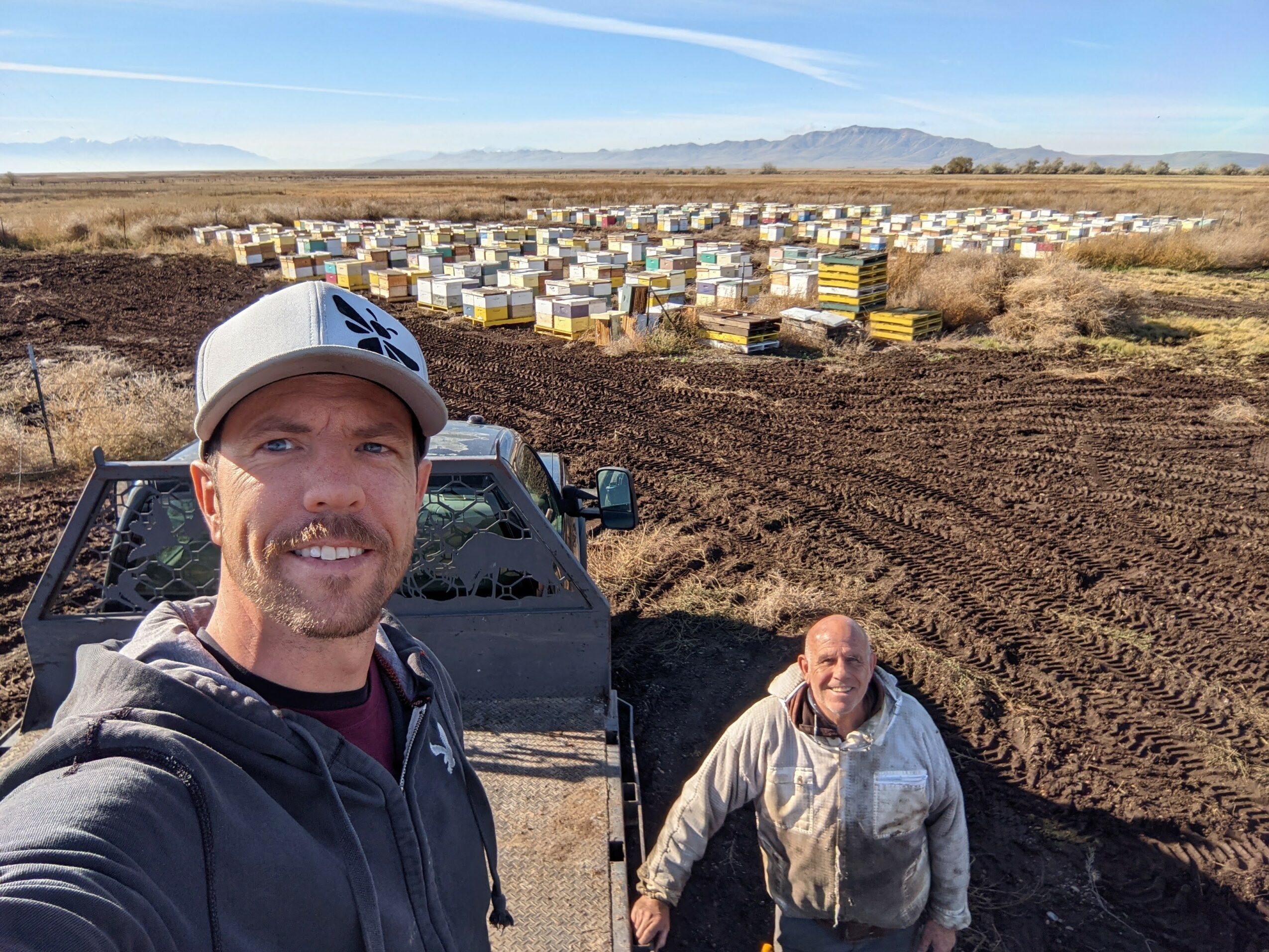 Two men outdoors in a field with a vehicle, one taking a selfie, and a large number of beehives in the background.
