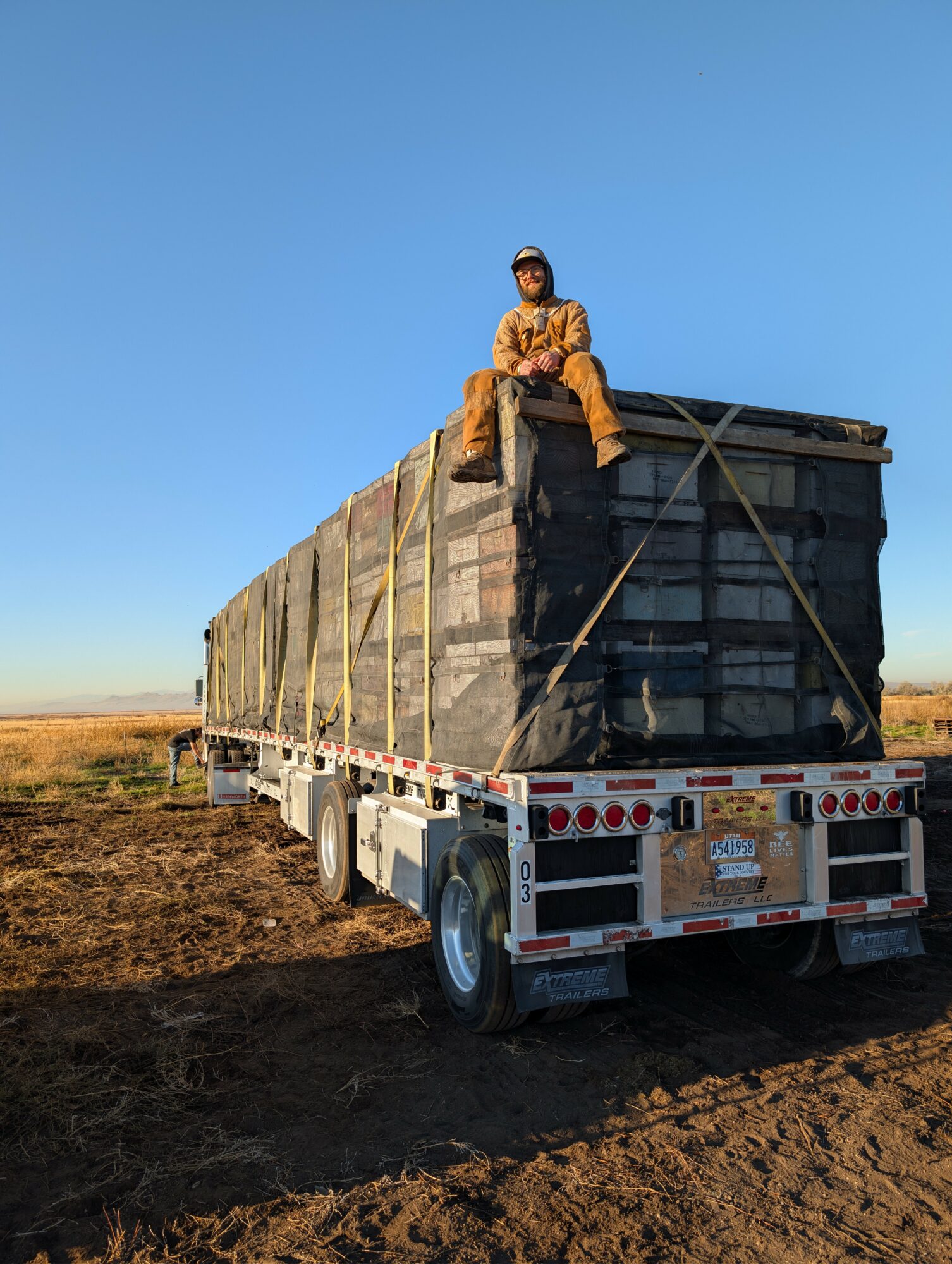Person sitting on a large truck loaded with black bags in an open field under a clear blue sky.