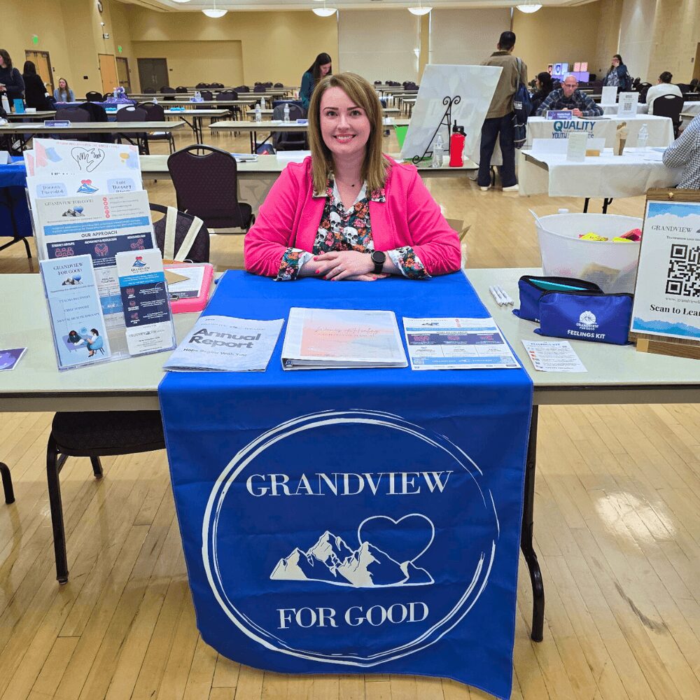 Woman sitting at a table with a blue tablecloth displaying 'Grandview for Good' logo, in a large room with tables and people.