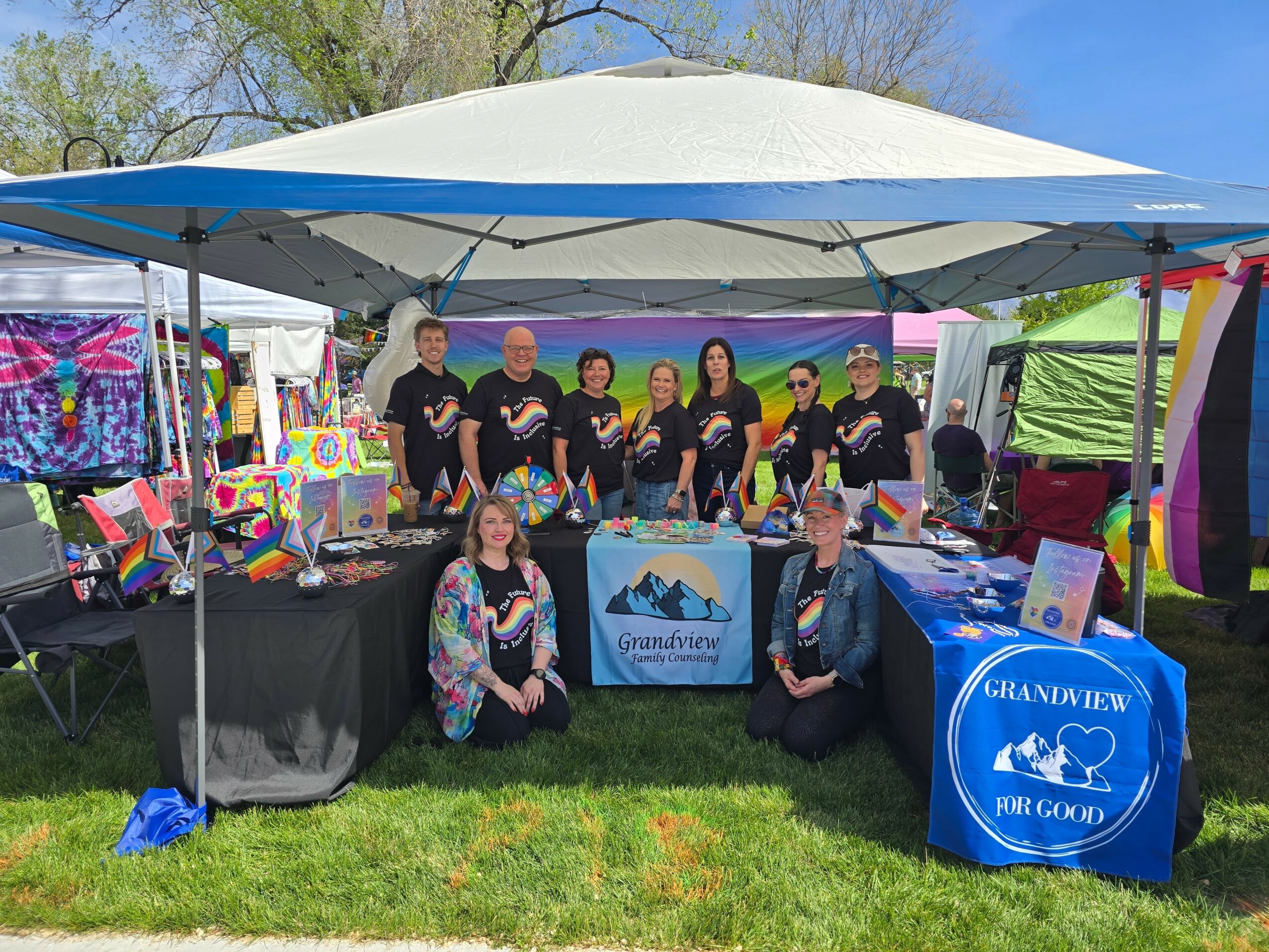 Group of people standing and sitting under a canopy at an outdoor event with tables displaying colorful items and banners.