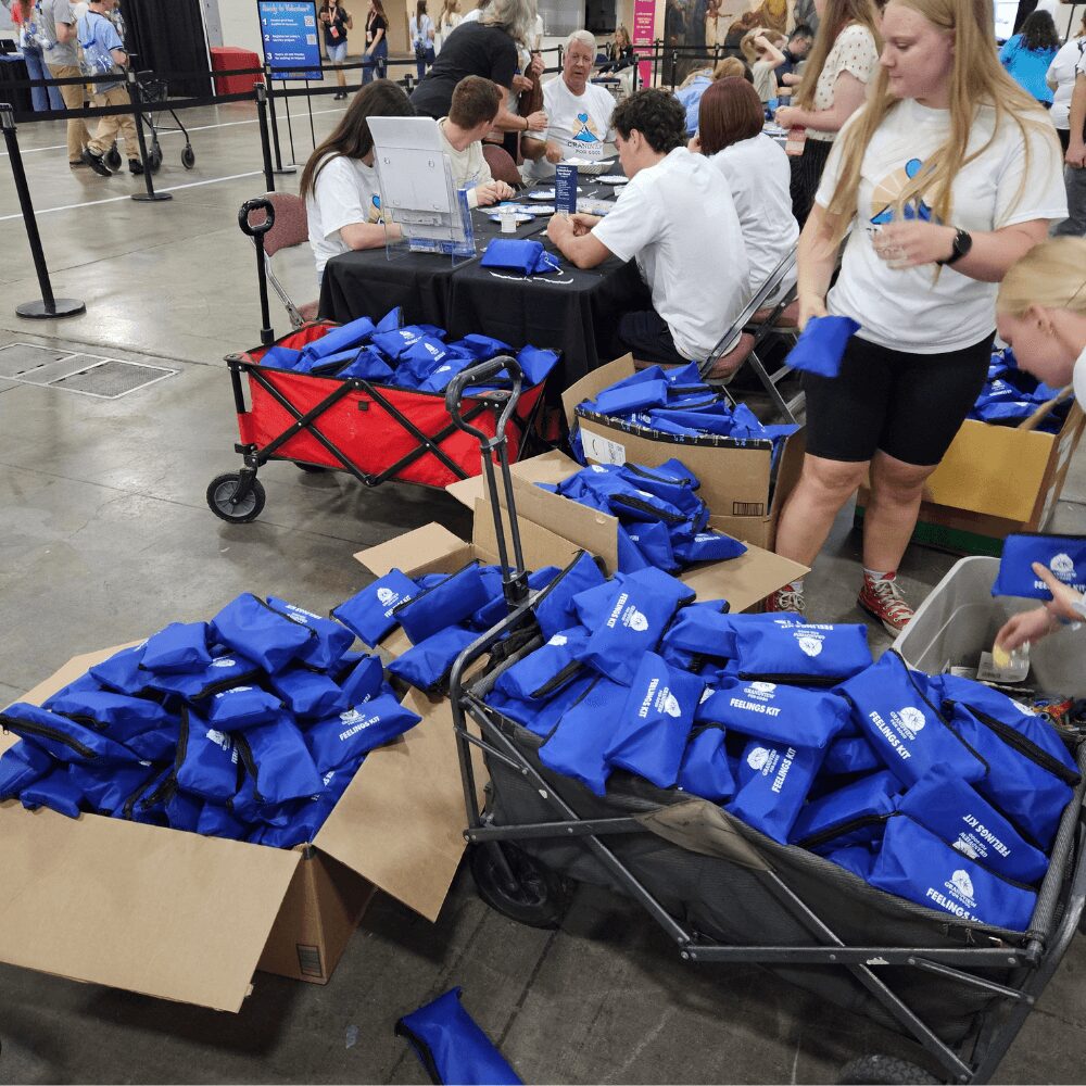 People gather around tables with blue bags and boxes in a large indoor space, possibly a registration or distribution area.