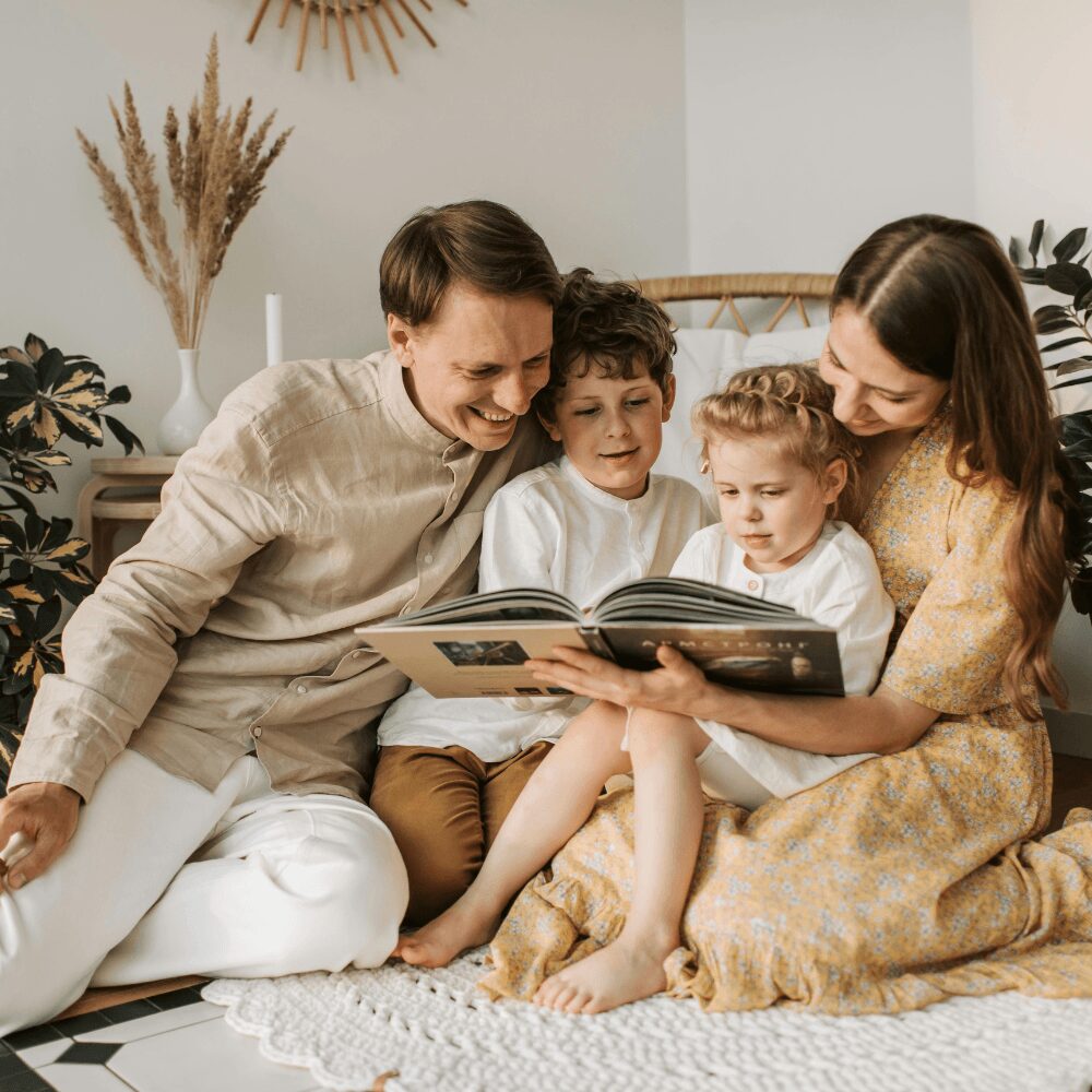 Family of four reading a book together on a white rug in a cozy room with plants and decor.