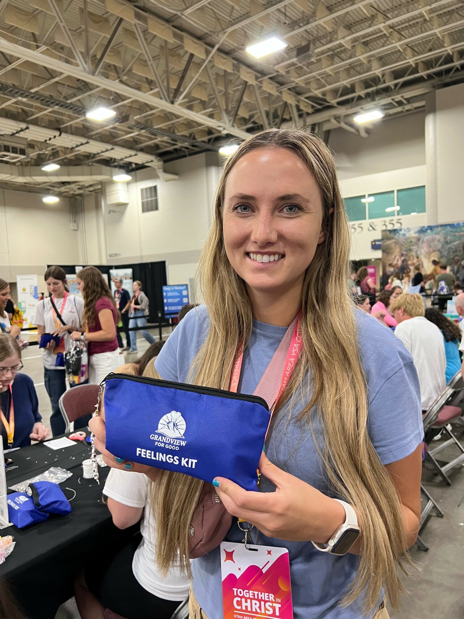 Young woman with long hair smiling, holding a blue Feelings Kit bag at an indoor event with people in the background.