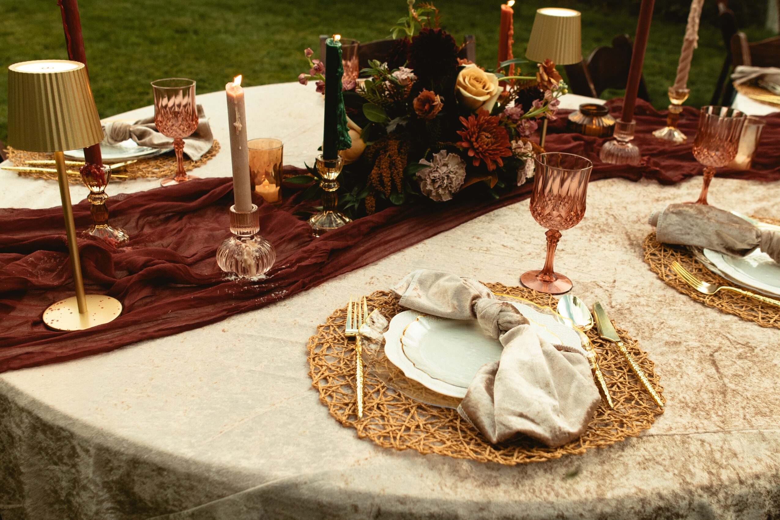 Elegant outdoor table setting with plates, glasses, candles, and floral centerpiece, on a cream-colored tablecloth with a burgundy runner.