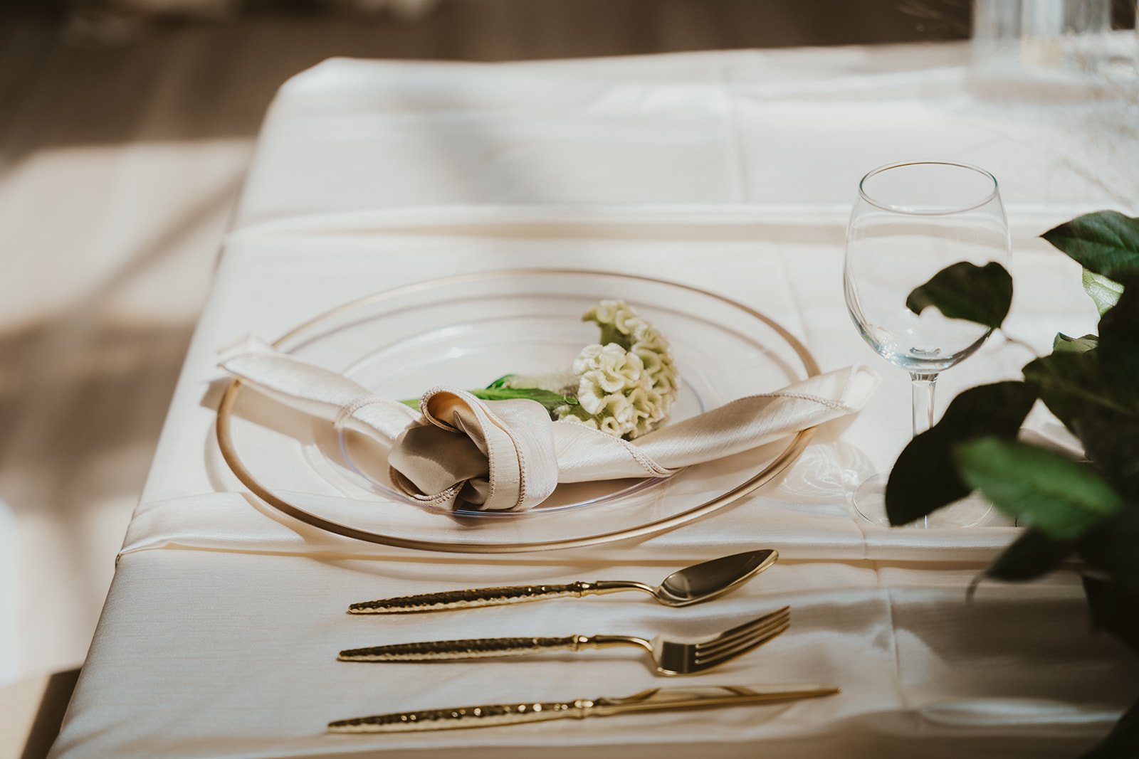 Elegant table setting with a white napkin, small flower, and gold cutlery on a white tablecloth, with a glass and greenery nearby.